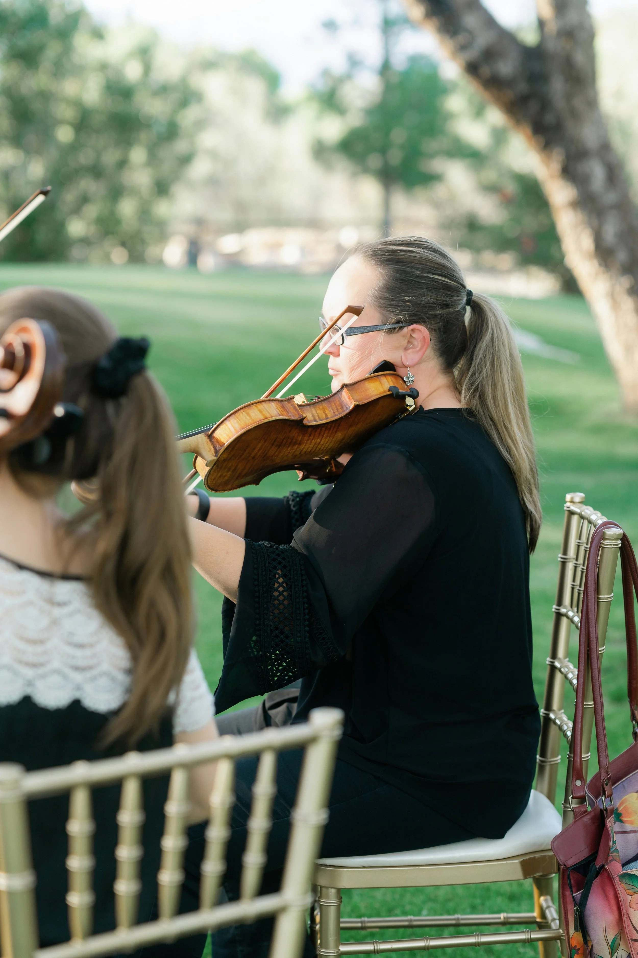 A woman with glasses and long hair playing the violin outdoors, seated in a gold-colored chair, with a green park and large tree in the background.