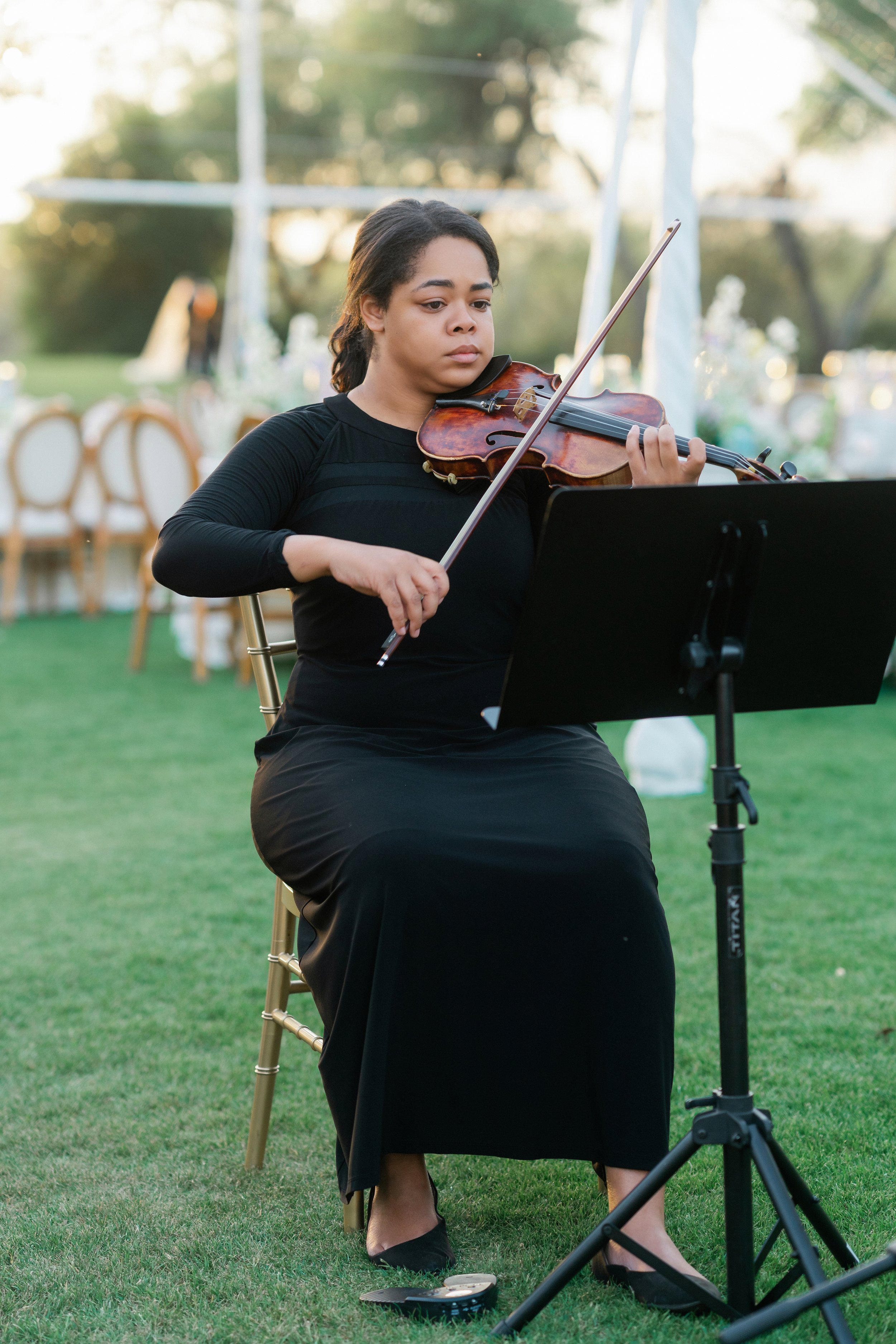 A woman in a black dress playing the violin outdoors at sunset, sitting on a wooden chair with a music stand in front of her, and an empty chair with a white cushion behind her.