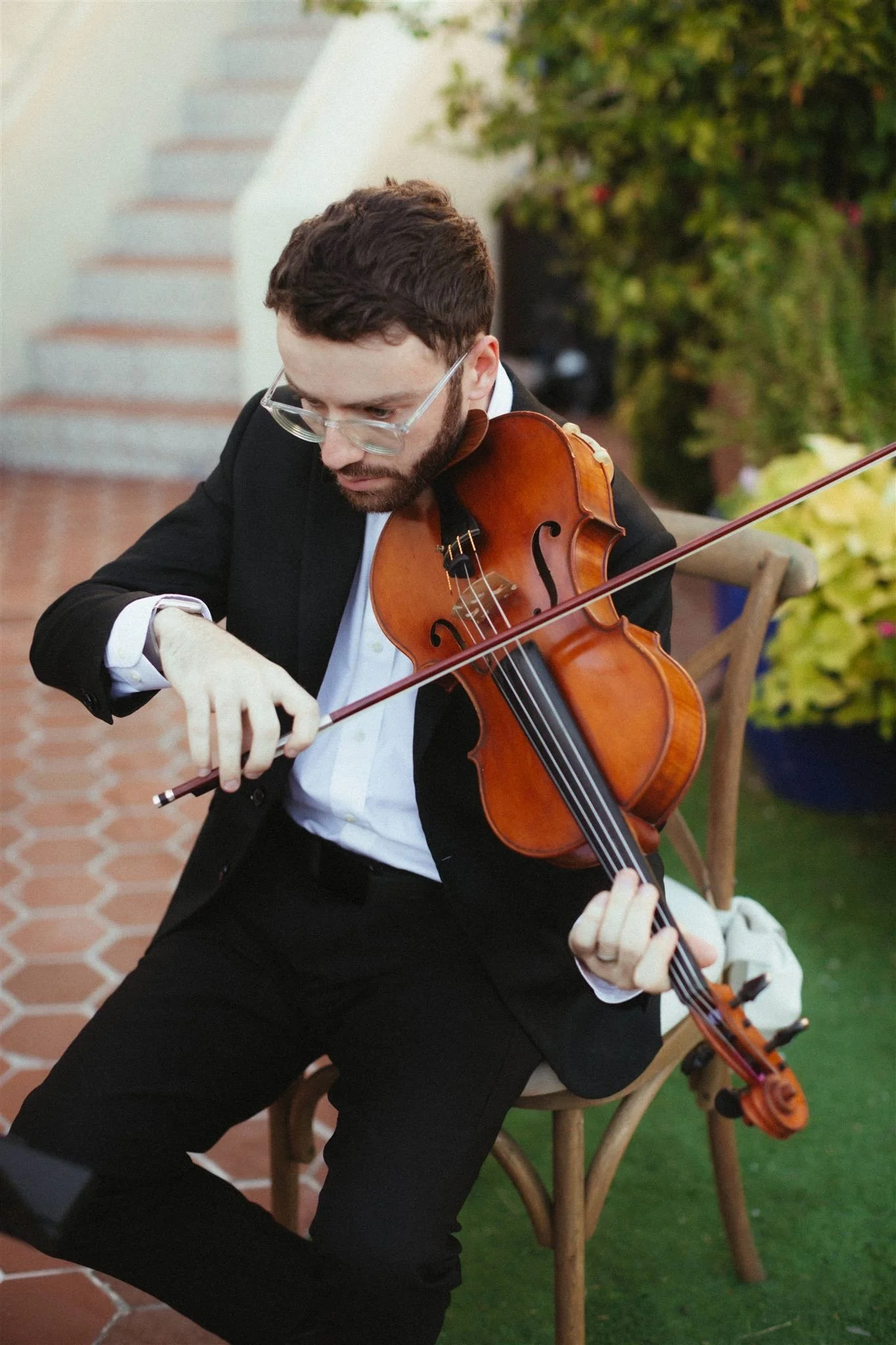 A man with dark hair and beard, wearing glasses and a black suit, playing the violin while sitting outdoors in a garden setting with green plants and a brick pathway.