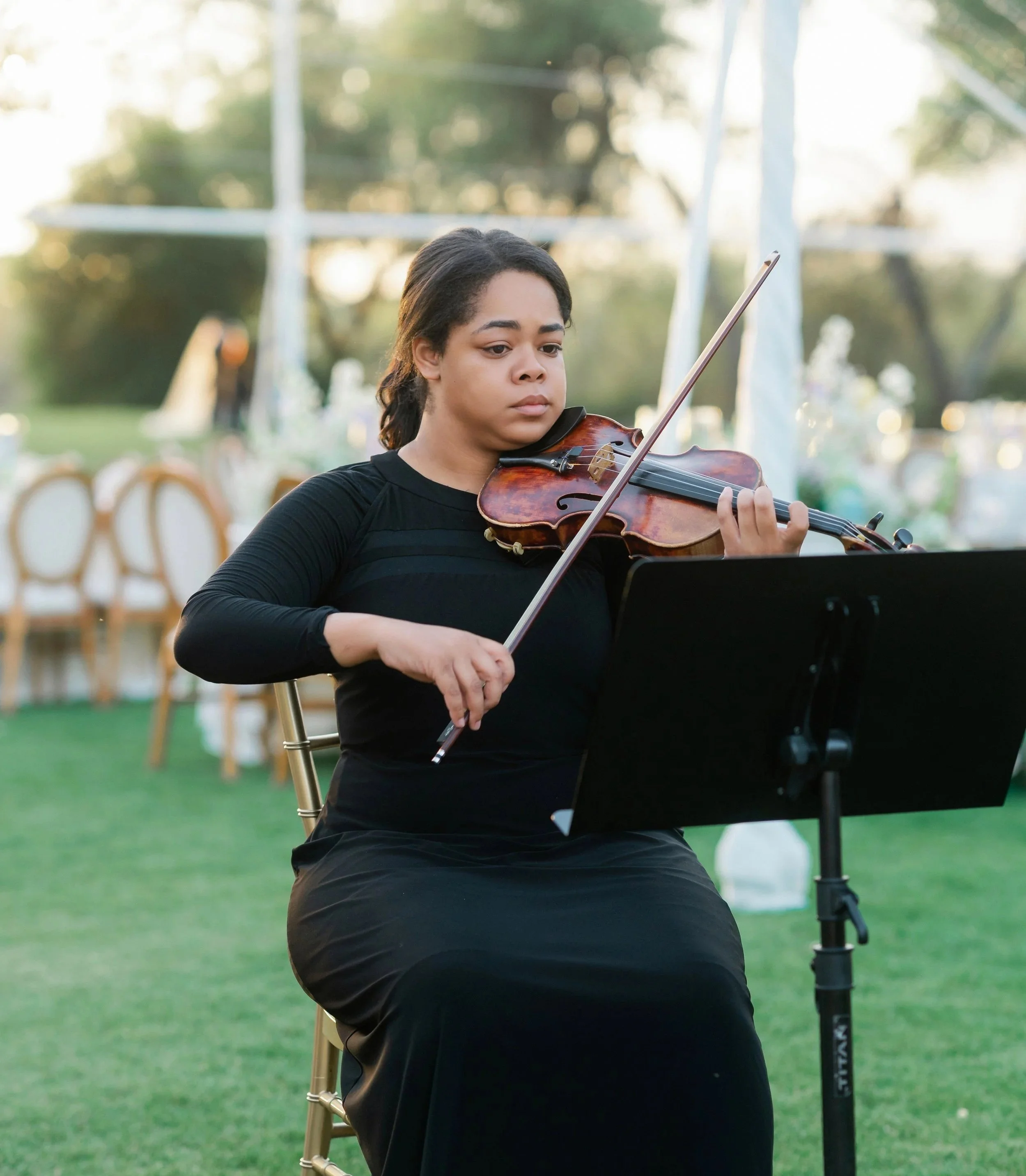 A woman playing the violin outdoors during sunset, with chairs and trees in the background.