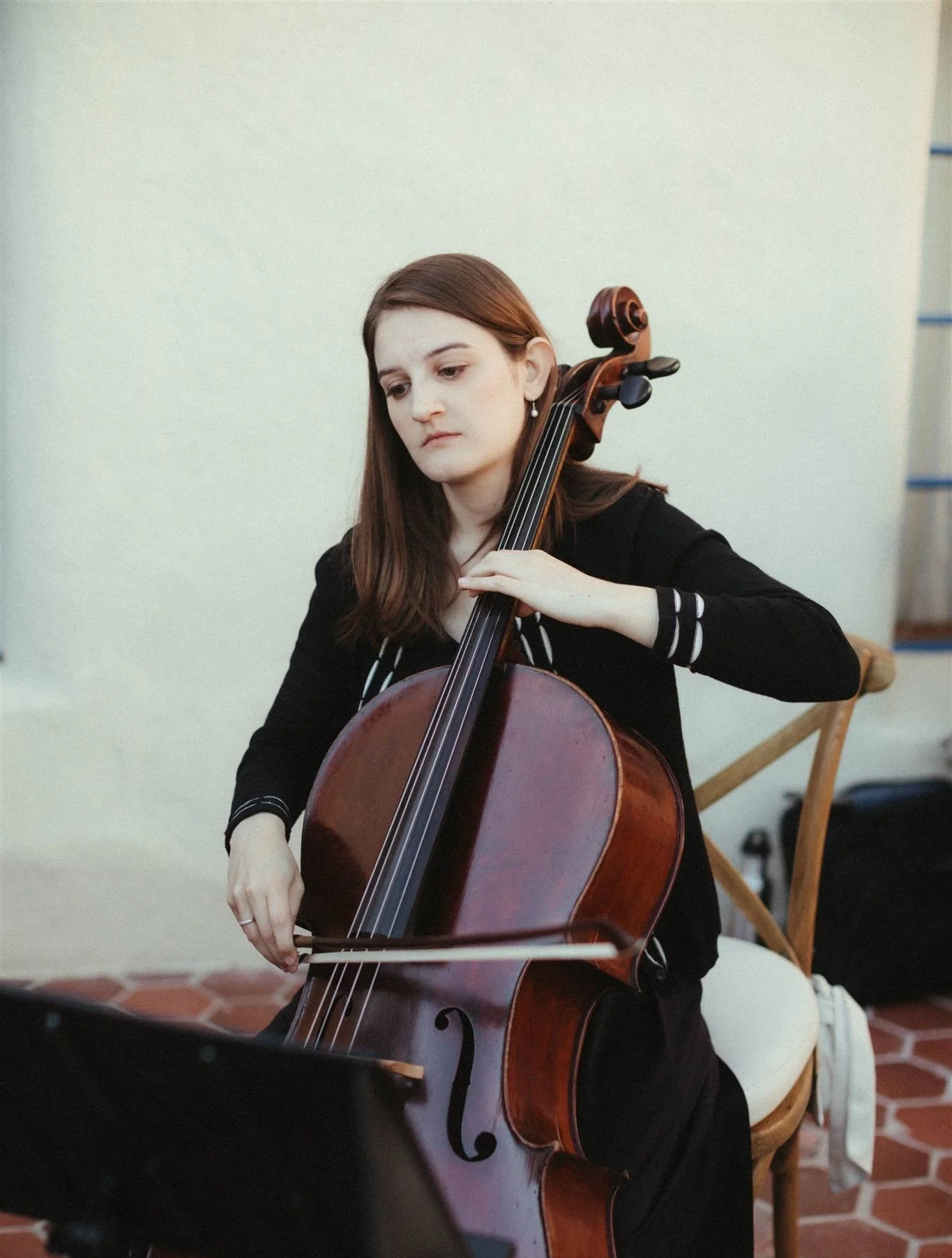 A young woman with long brown hair playing a cello indoors. She is wearing a black top with some white patterns on the sleeves and appears focused on her music. The background features a plain white wall and part of a wooden chair.