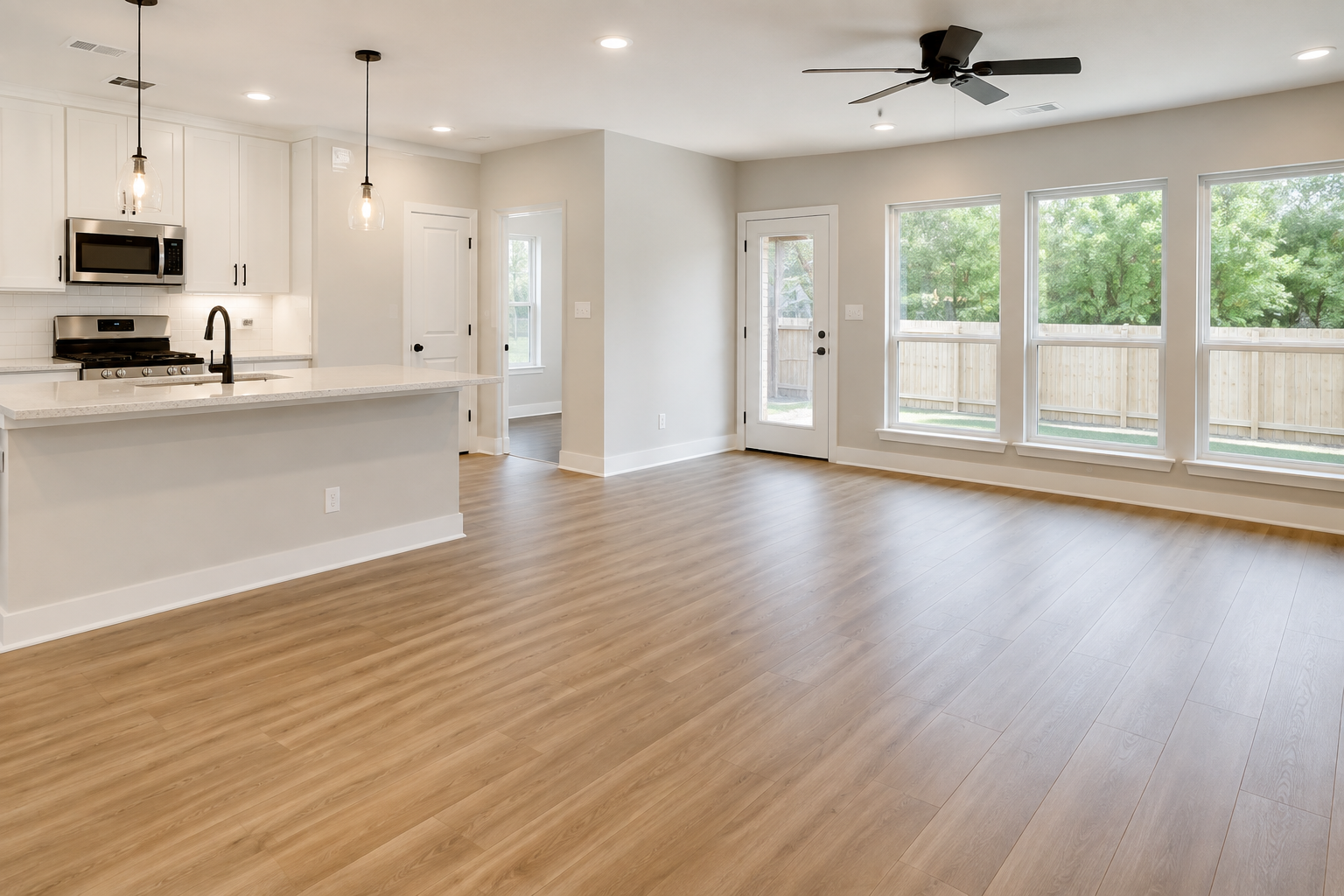 Empty living room area with light wood flooring, large windows, and a doorway leading outside. Part of the kitchen with white cabinets, a black faucet, and two hanging pendant lights.
