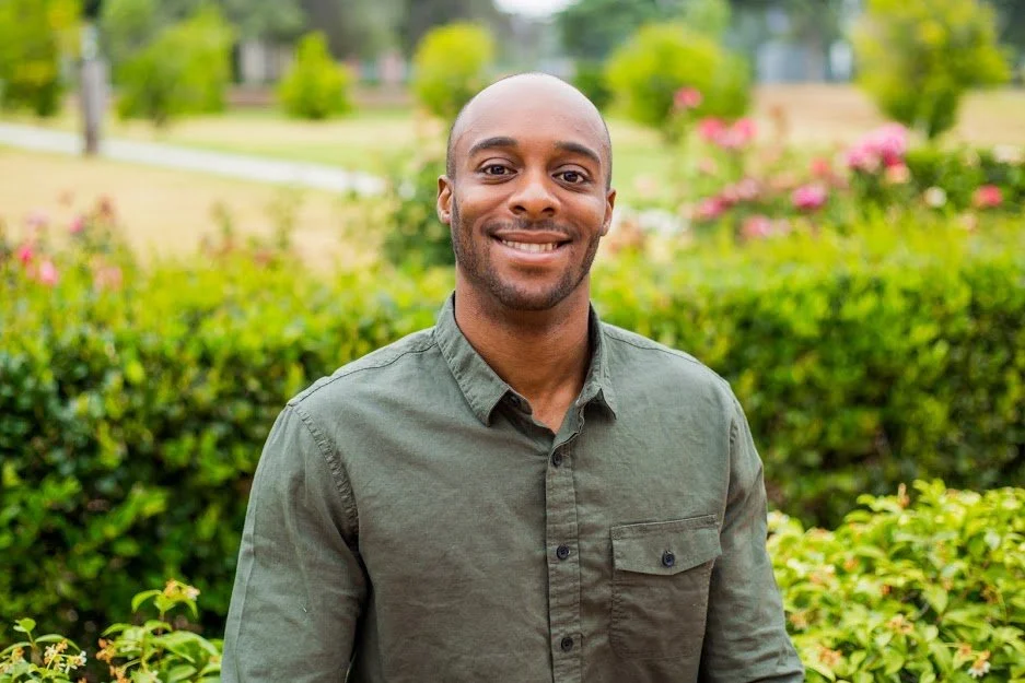 A smiling man standing outdoors in a park with green bushes and pink flowers in the background.