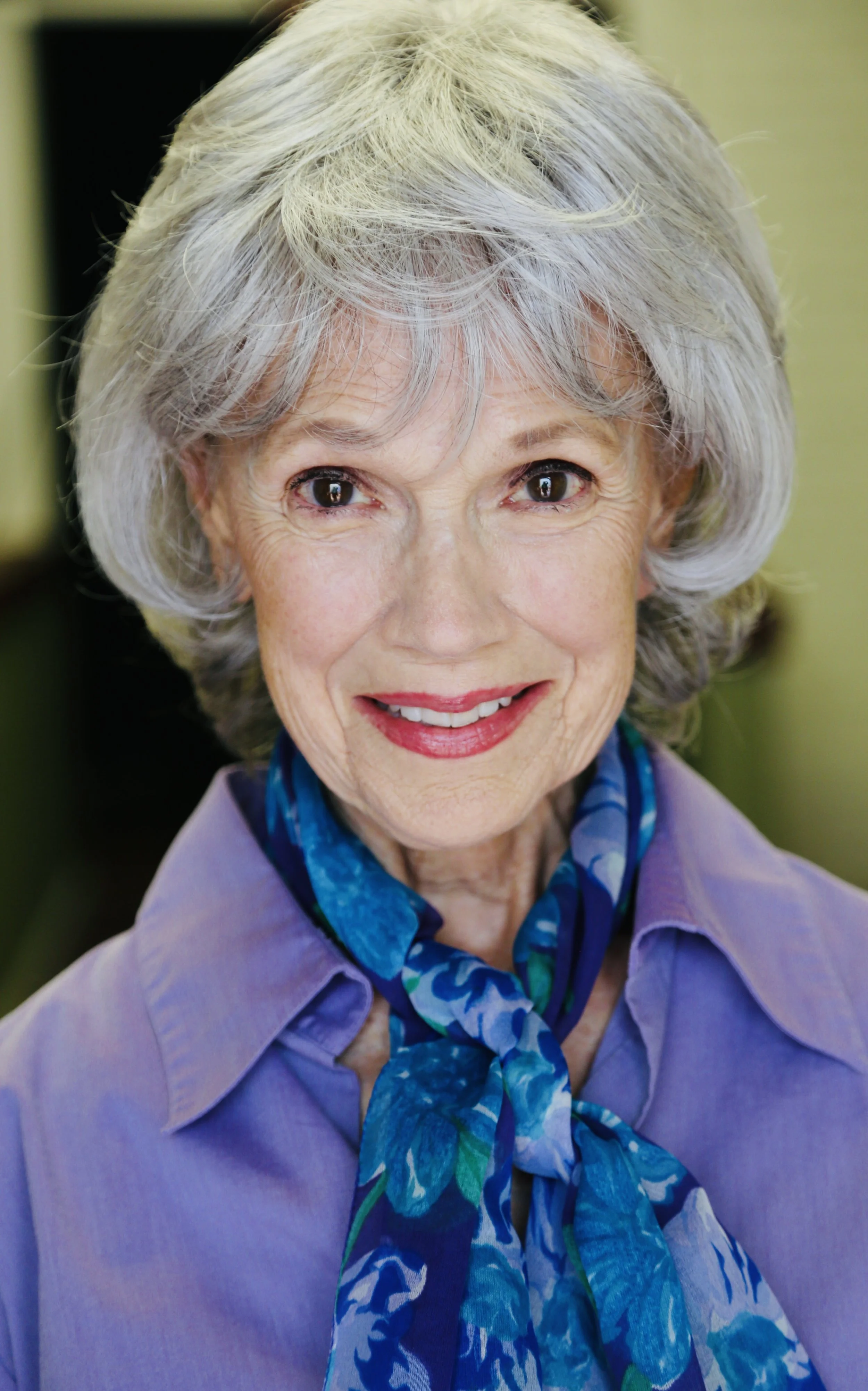 Close-up of Mary O' Kerrigan with gray hair, smiling and wearing a purple blouse with a blue patterned scarf.