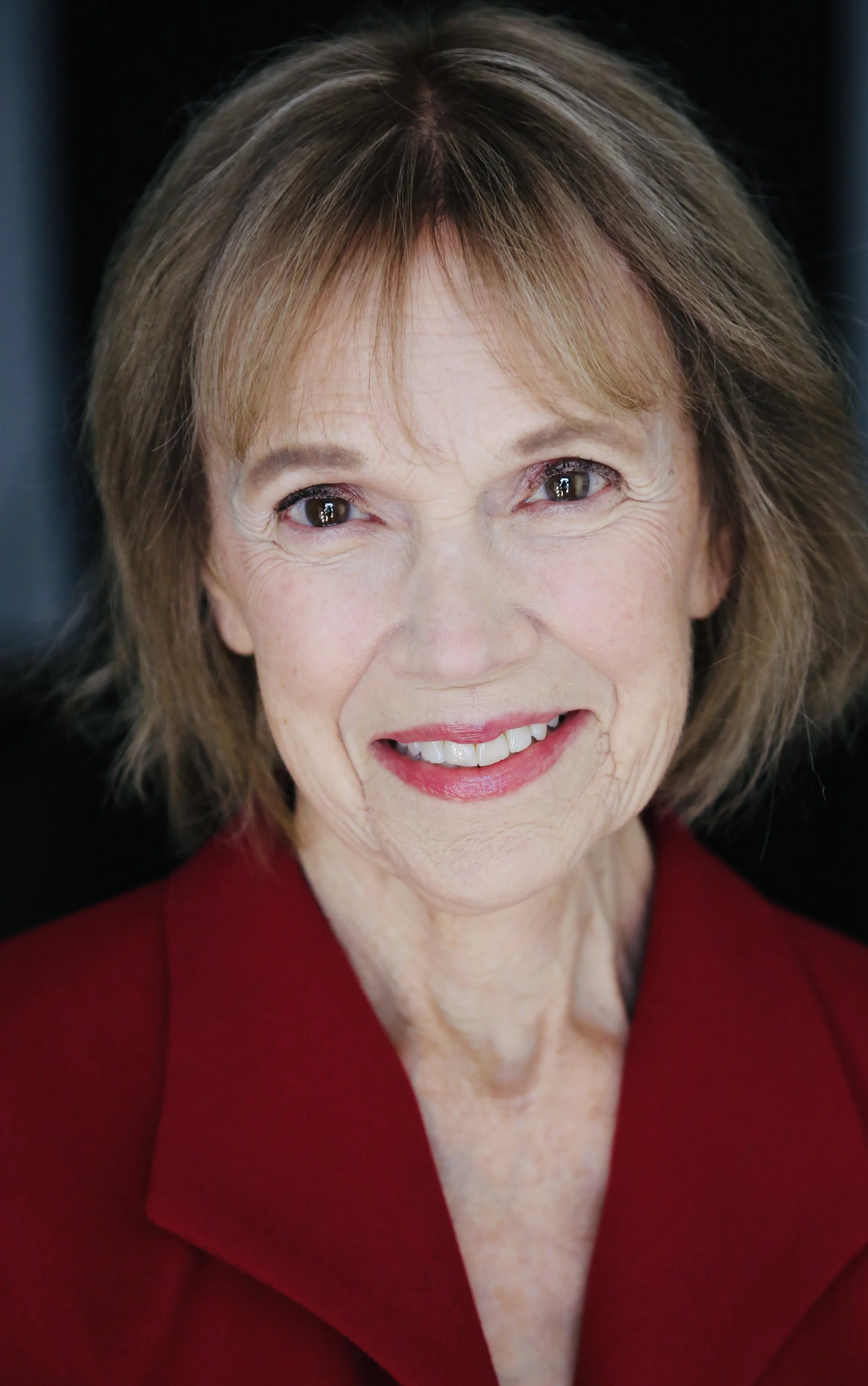 Close-up portrait of Mary O' Kerrigan with short, light brown hair and a bright smile, wearing a red blazer, against a dark background.