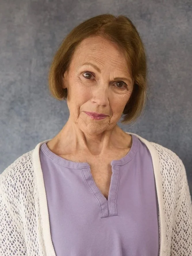 A portrait of an elderly woman with short brown hair, wearing a lavender top and cream-colored cardigan, standing against a gray background.