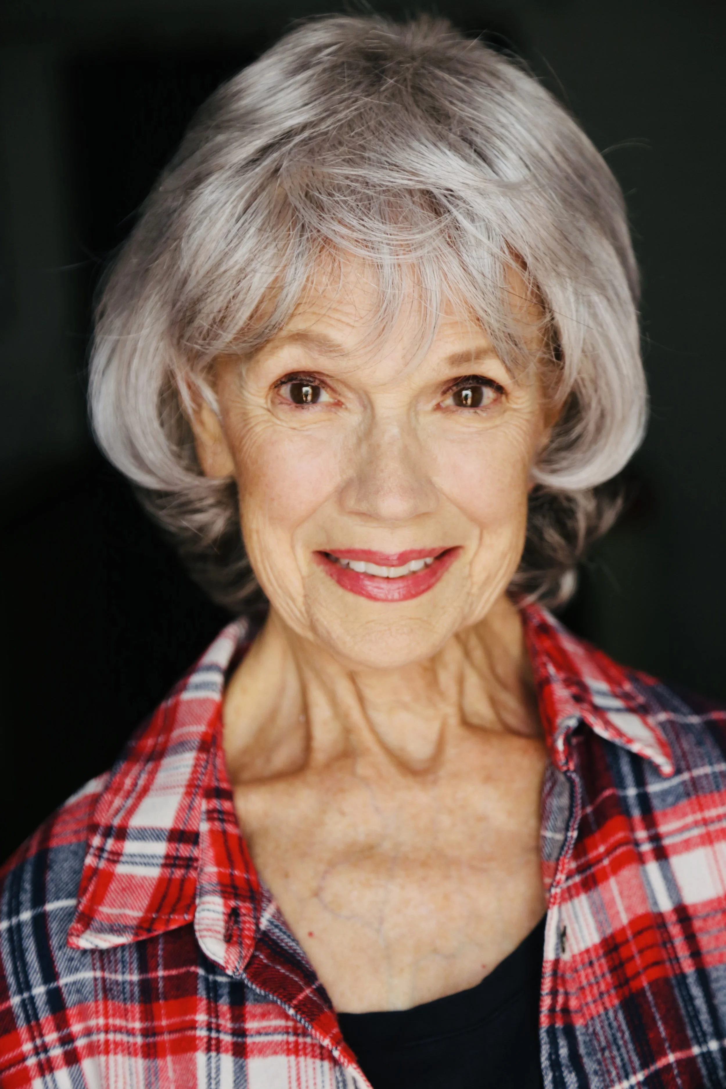 Close-up of Mary O' Kerrigan with short, wavy gray hair, wearing a red plaid shirt over a black top about to smile.