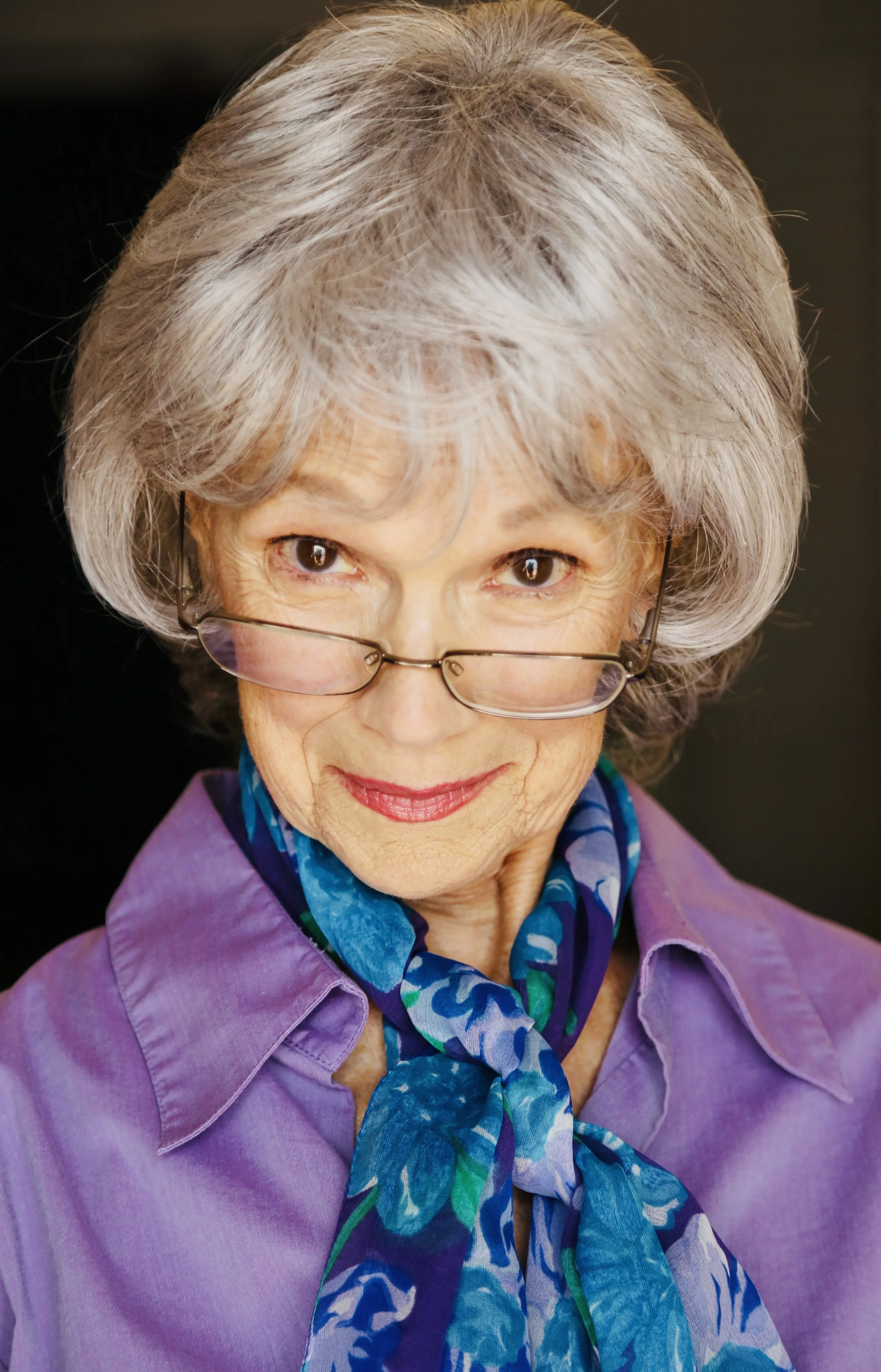 Close-up portrait of Mary O' Kerrigan with gray hair, wearing glasses, a purple shirt, and a patterned blue scarf, smiling slightly.