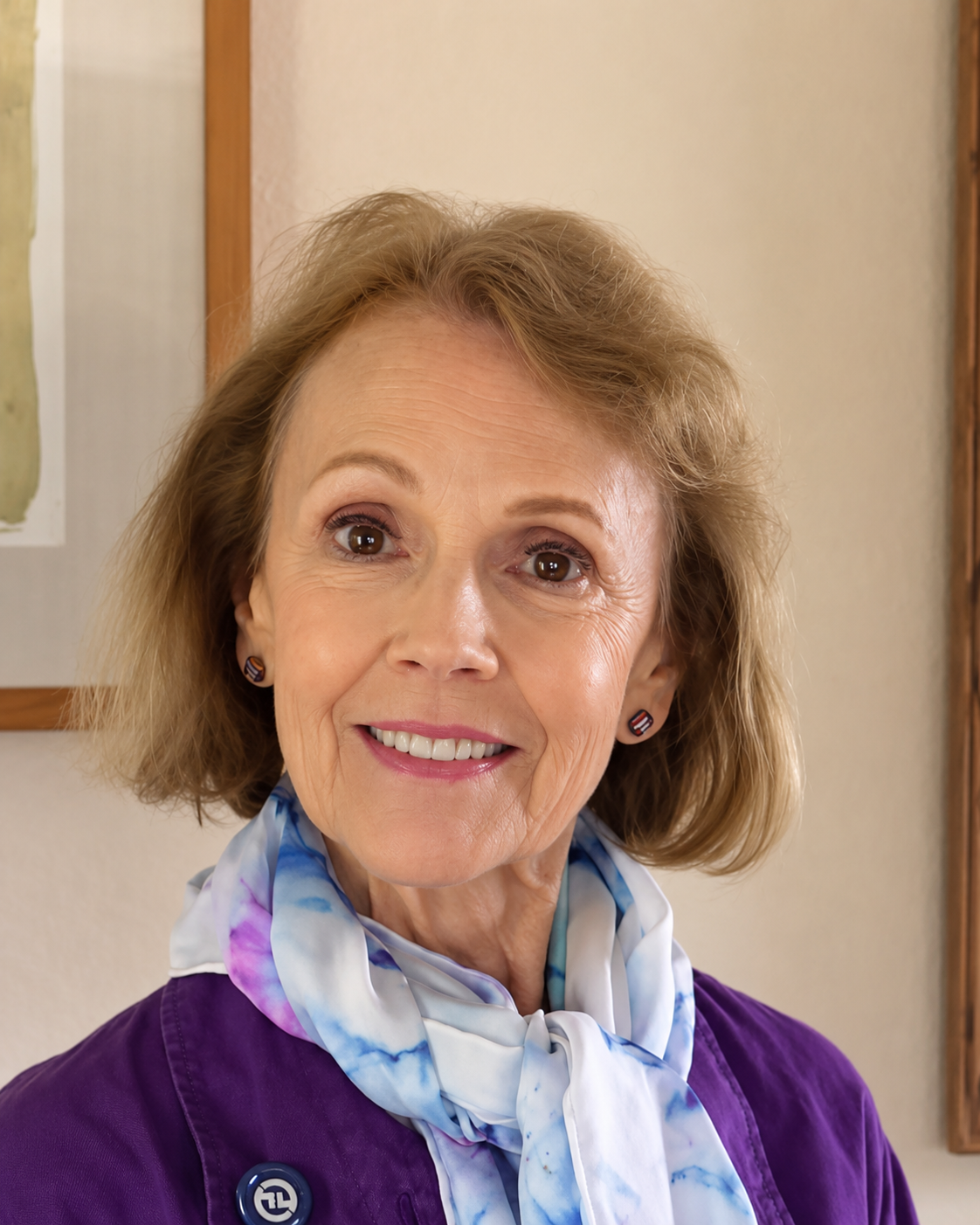 Close-up of Mary O' Kerrigan with light brown hair smiling, wearing a purple jacket and a white-blue floral scarf, in a room with framed artwork on the wall.