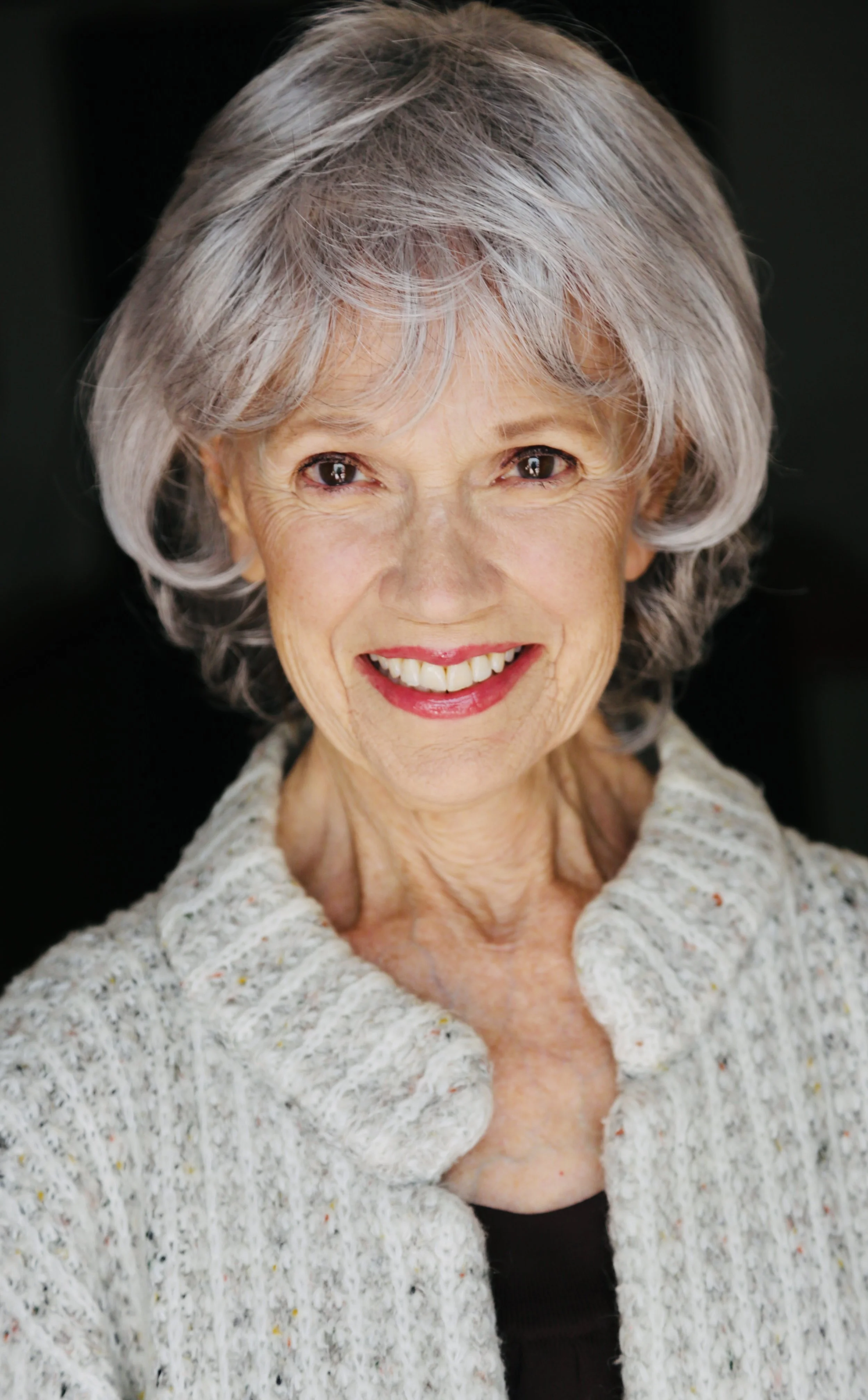 Portrait of Mary O' Kerrigan with short gray hair, smiling, wearing a light-colored, speckled cardigan and a black top, against a dark background.