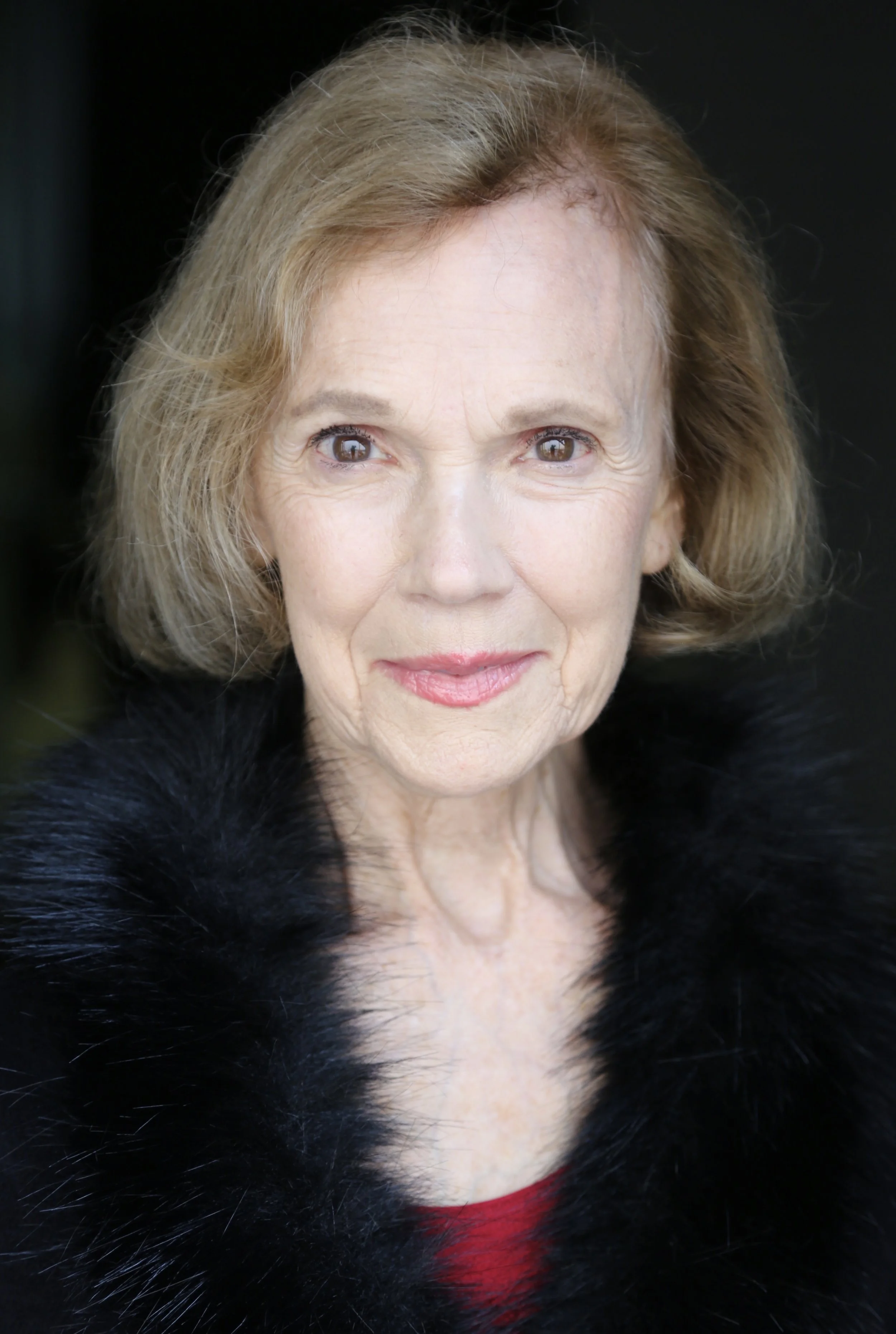 Close-up of Mary O' Kerrigan with short blonde hair, wearing a black fur coat, standing against a dark background.