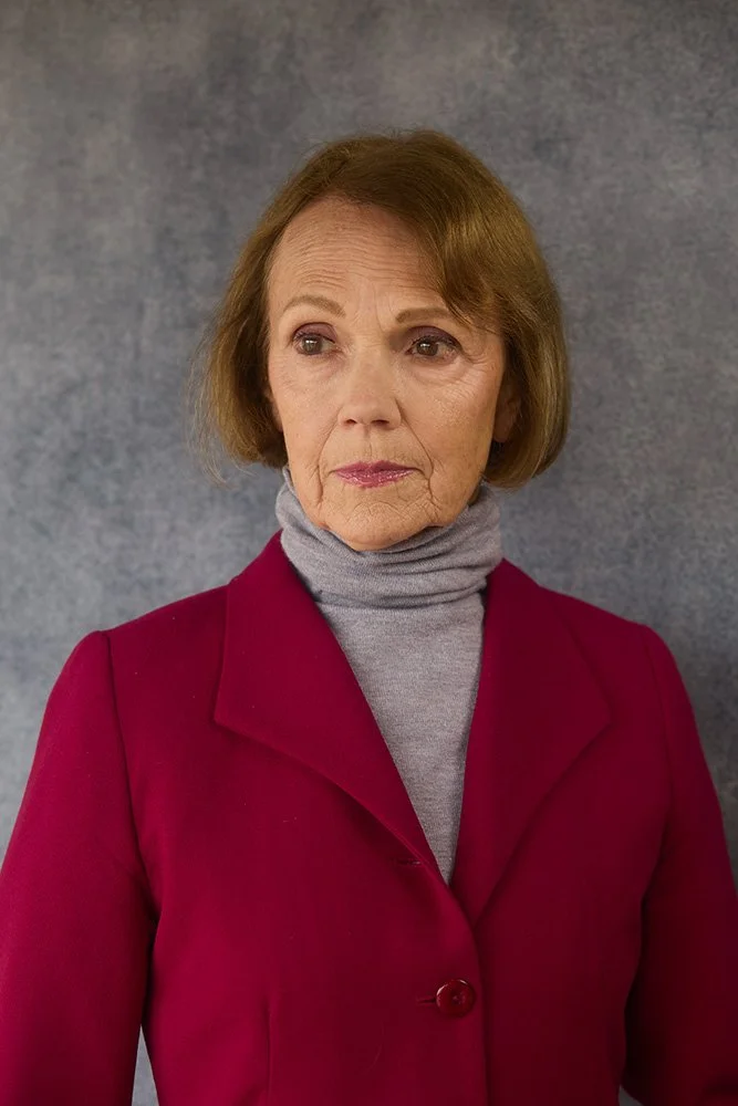 Mary O' Kerrigan with short light brown hair wearing a gray turtleneck and a burgundy blazer stands against a plain gray background, looking to the side with a neutral expression.
