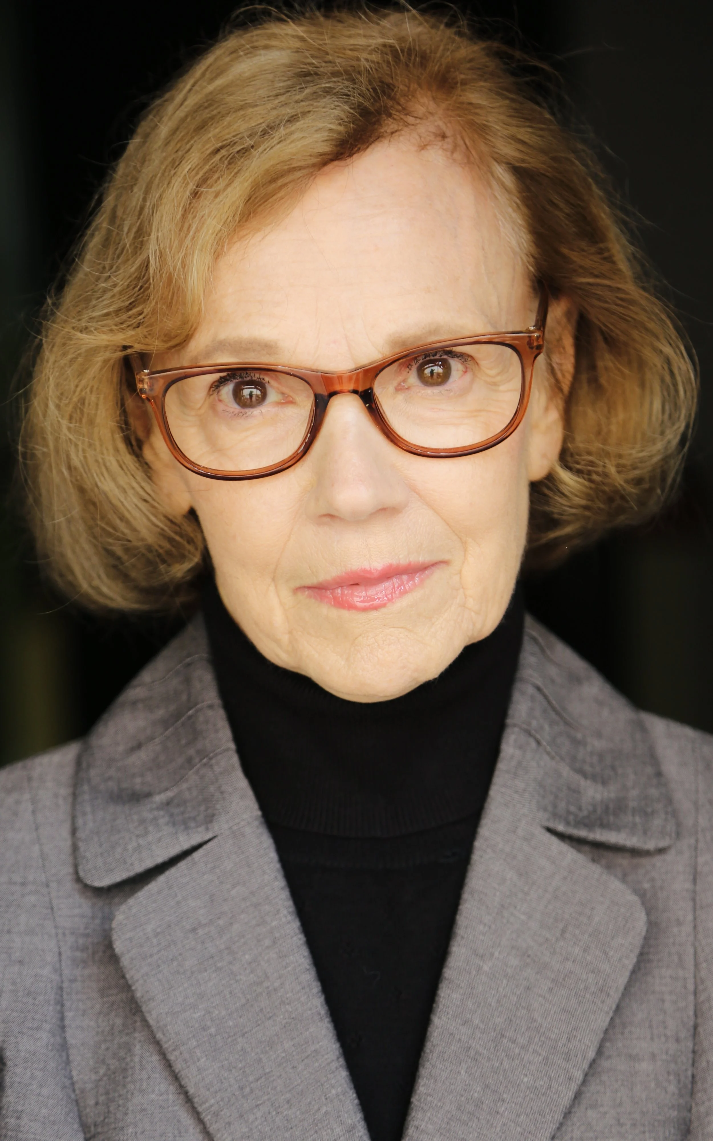 Close-up of Mary O' Kerrigan with short light brown hair, wearing brown-framed glasses, a black turtleneck, and a gray blazer with a subtle checkered pattern.