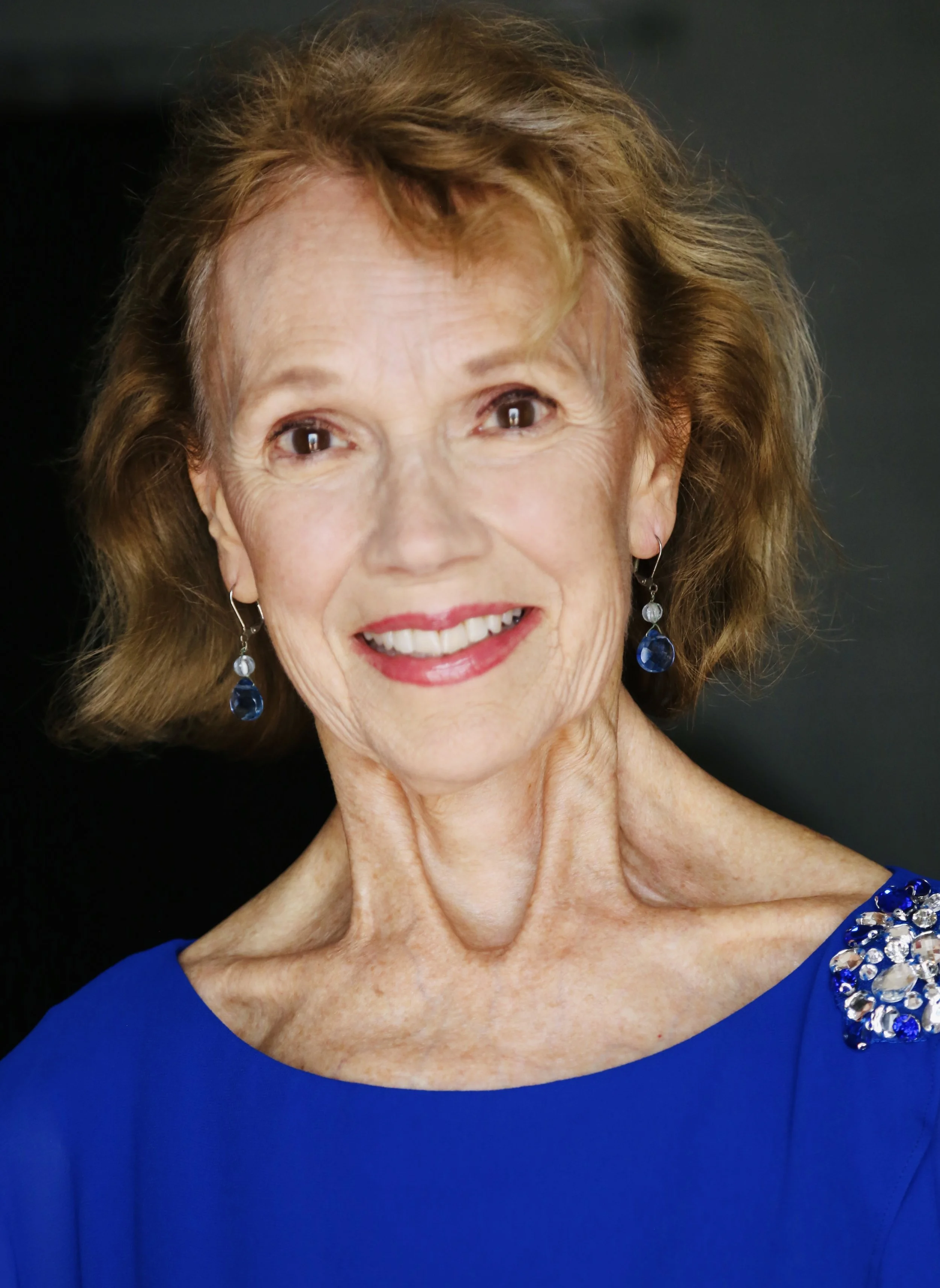 Close-up of Mary O' Kerrigan with curly auburn hair smiling, wearing blue earrings with blue gemstone accents and a royal blue dress with embellished shoulder detail, posed against a dark background.