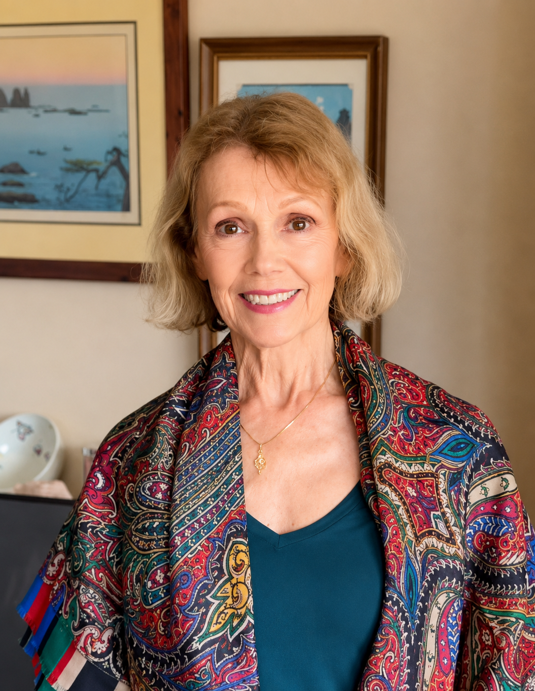 Mary O' Kerrigan with blonde hair smiling at the camera, wearing a colorful patterned jacket and a teal top, standing in a room with framed artwork on the wall.
