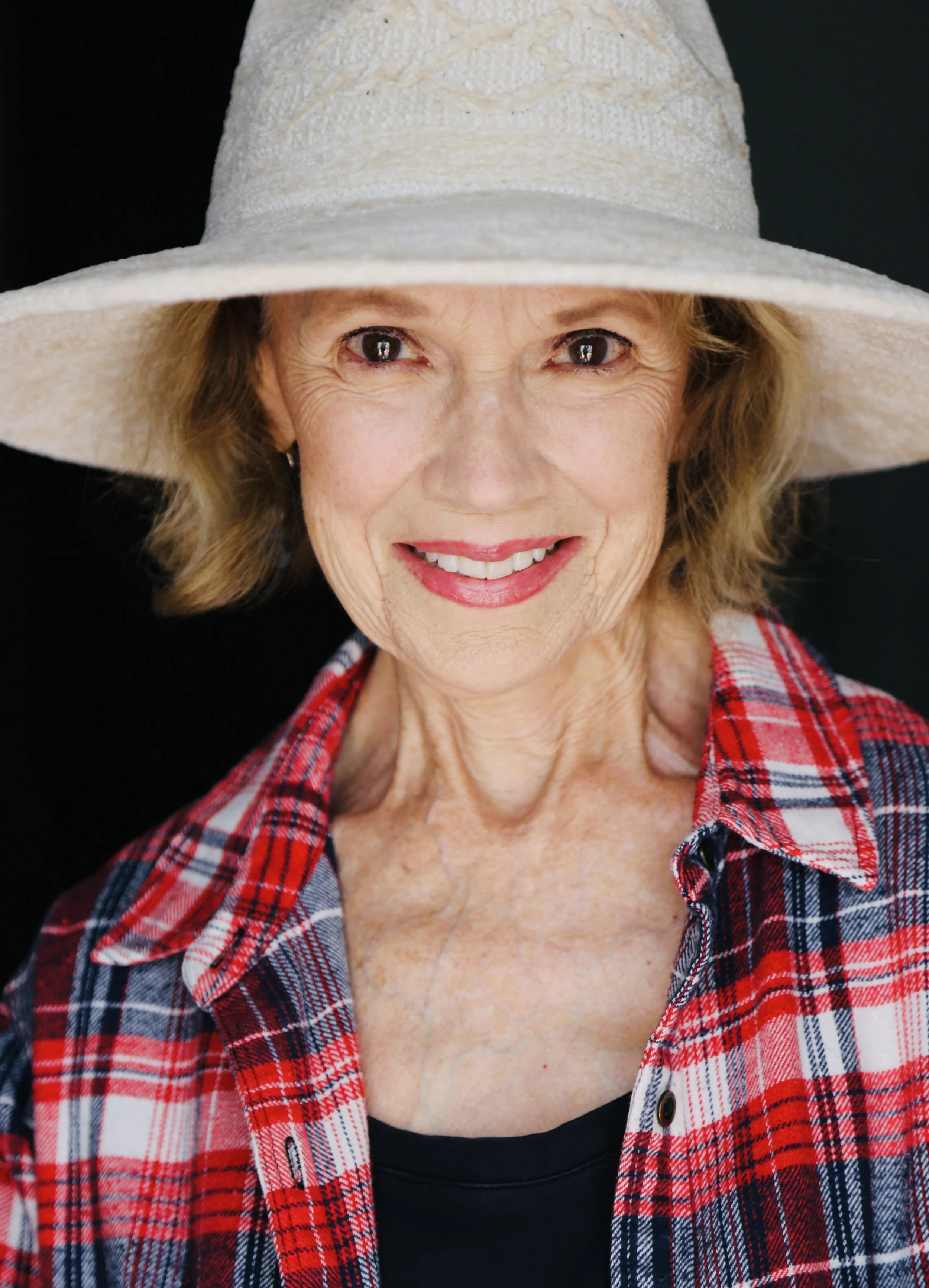 Close-up of Mary O' Kerrigan wearing a beige wide-brimmed hat, smiling and dressed in a red plaid shirt over a black top, against a black background.