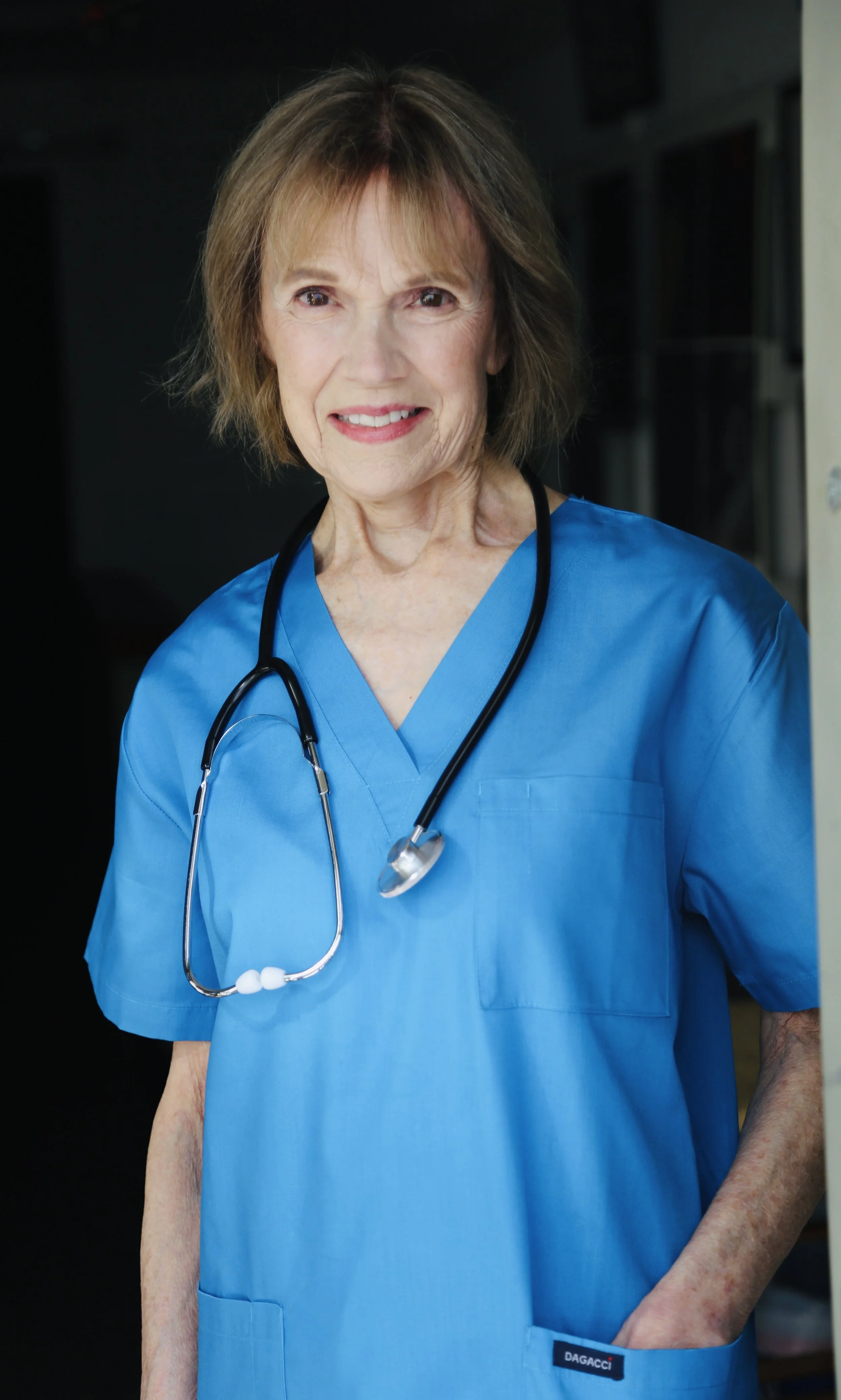 Close-up of a senior woman wearing blue medical scrubs and a stethoscope, standing outdoors with a blurred background.