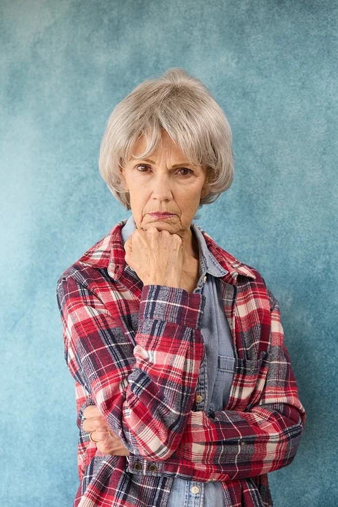 An older woman with gray hair, wearing a red plaid shirt over a denim shirt, standing with her arm crossed and her hand resting on her chin against a blue background.