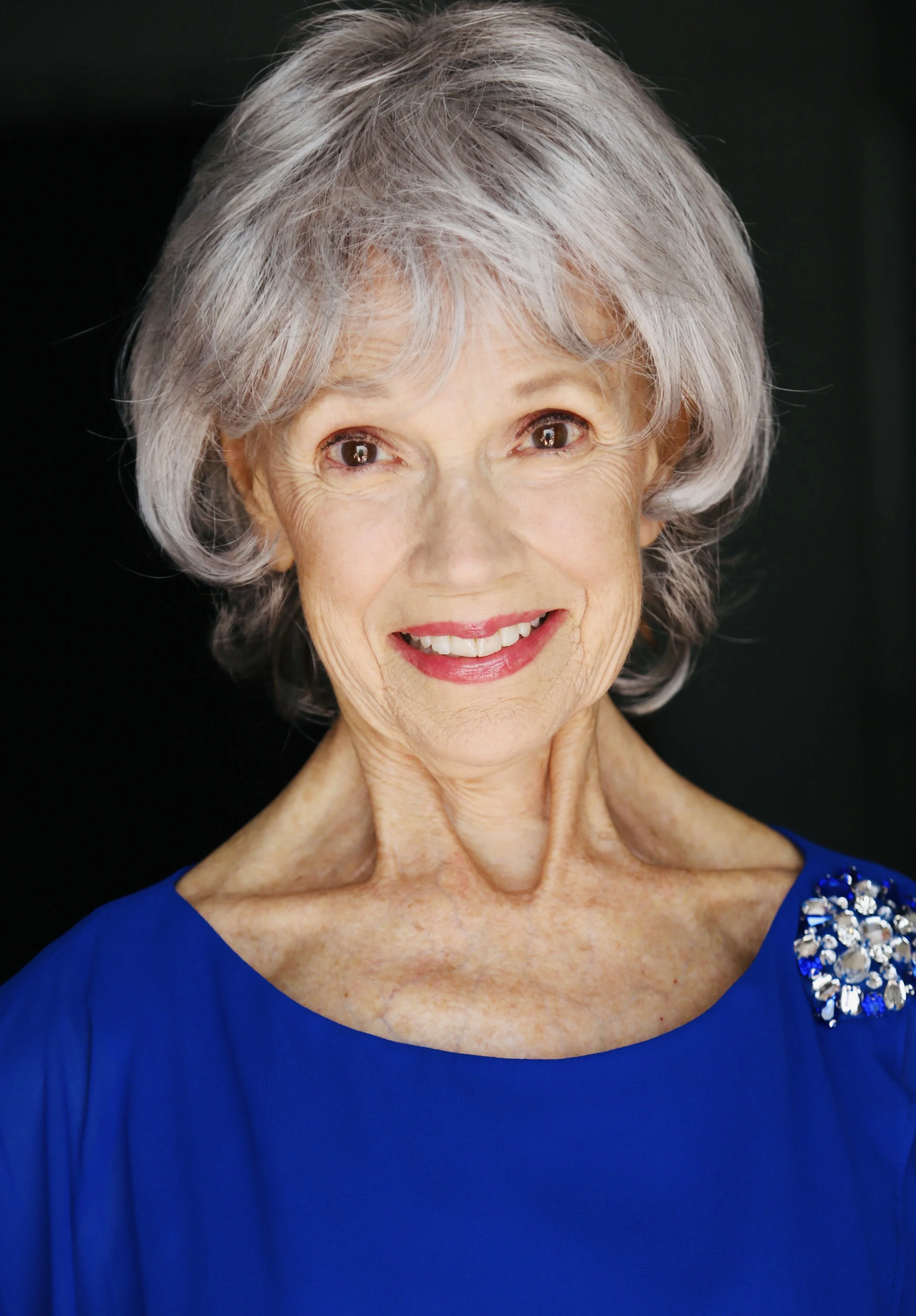 Close-up of an elderly woman with short gray hair, smiling, wearing a royal blue dress with embellishments on the shoulder, against a black background.