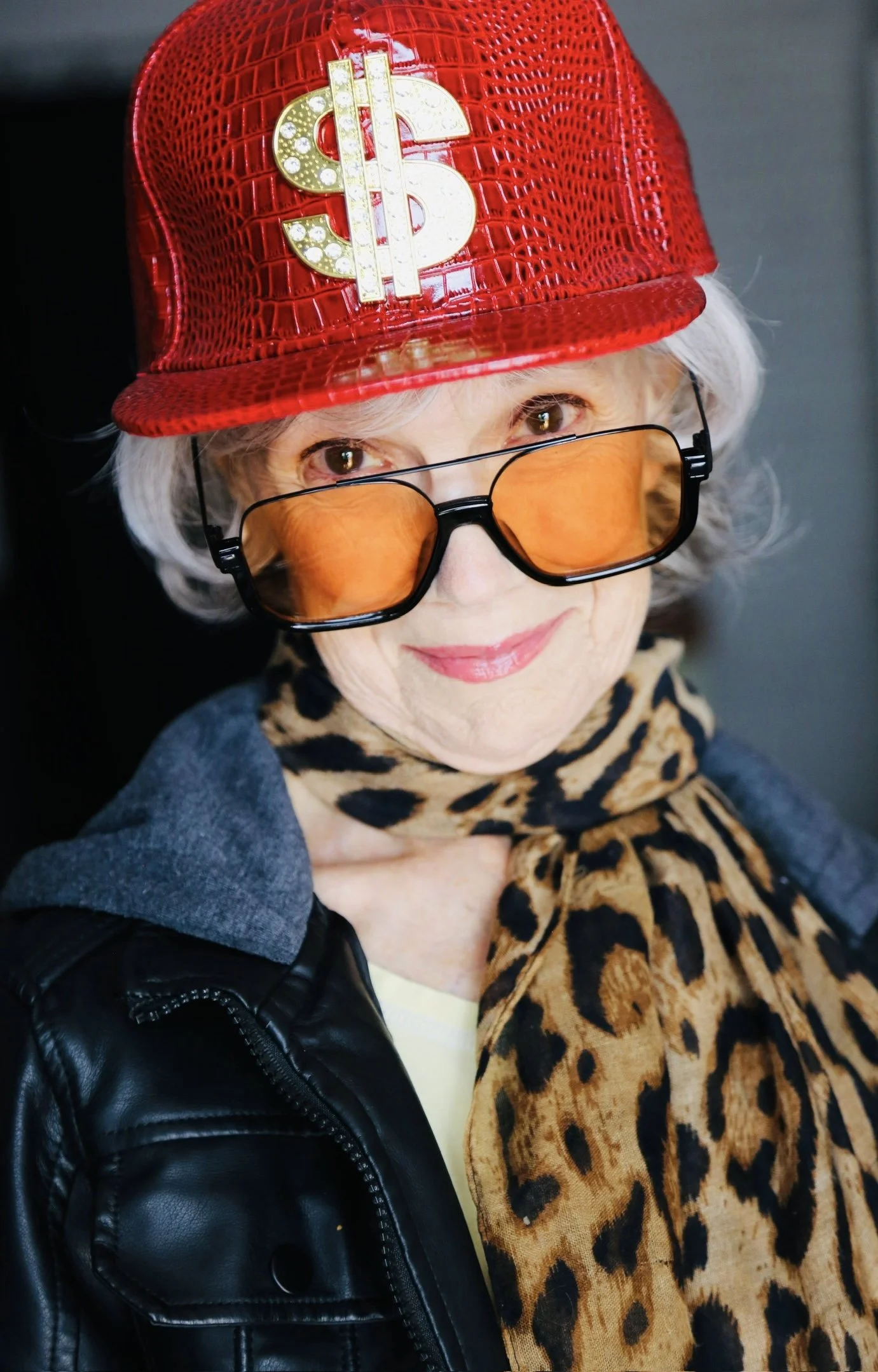 Close-up portrait of Mary O' Kerrigan wearing a red crocodile-patterned baseball cap with a gold dollar sign pin, orange-tinted sunglasses, a leopard print scarf, and a black leather jacket.