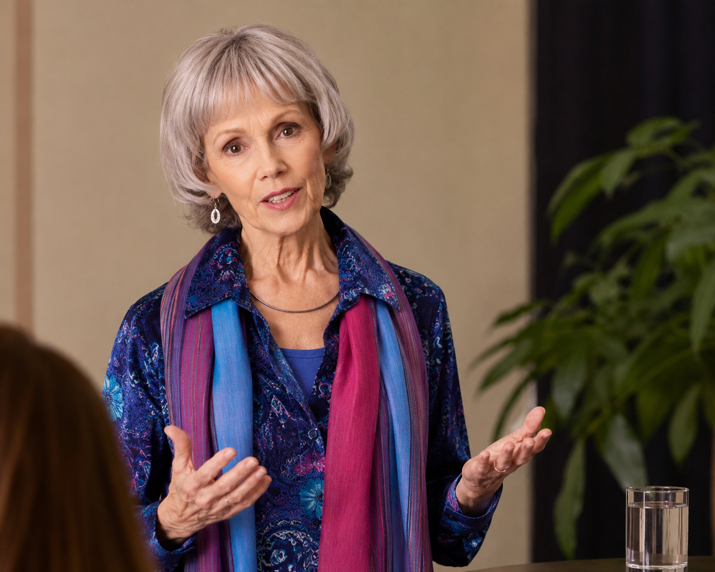 Mary O' Kerrigan speaking, wearing a blue floral shirt with a colorful scarf, in a room with beige walls, a large green plant, and a glass of water on the table.