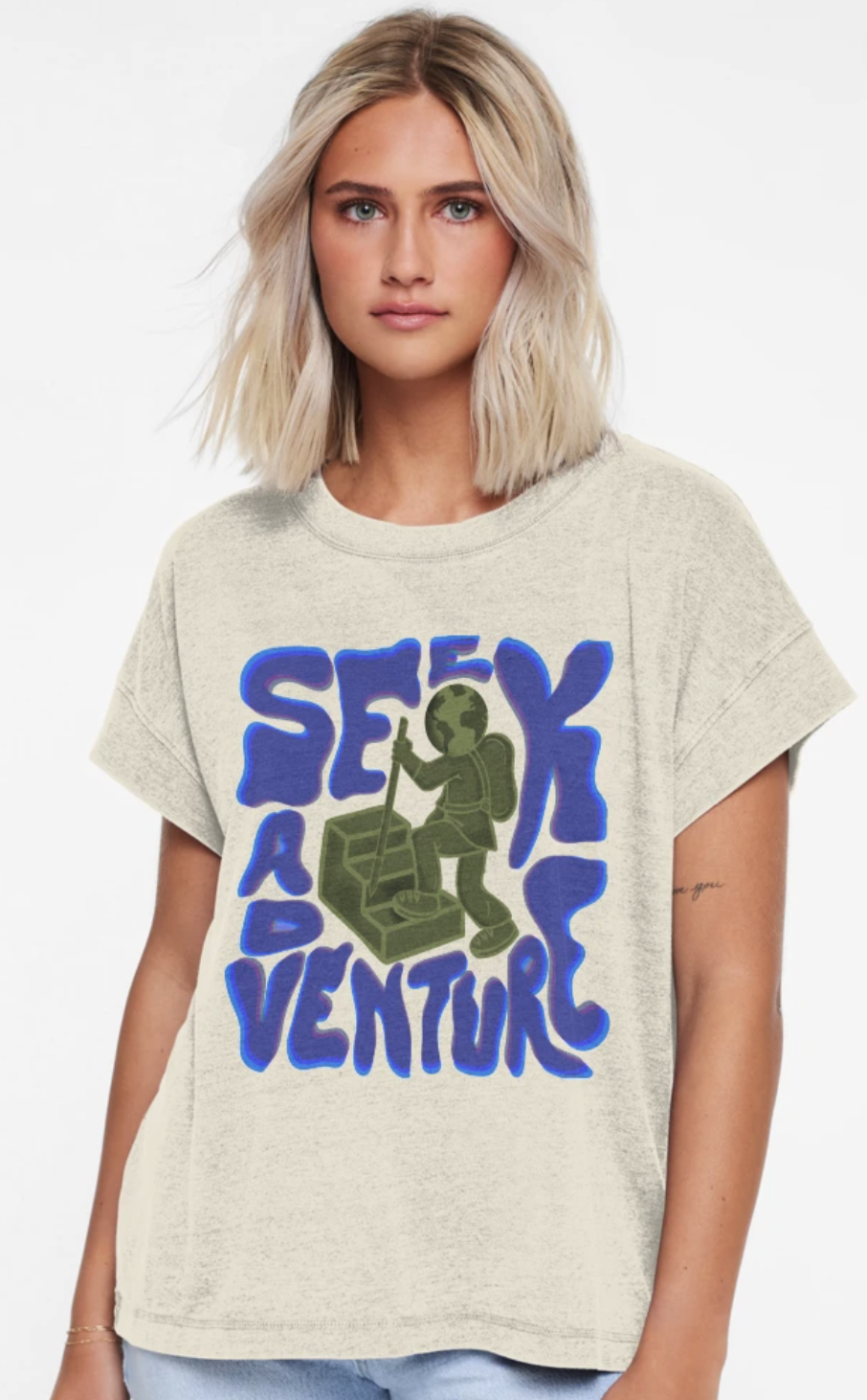 A young woman with blonde hair wearing a beige T-shirt with a green graphic of a hiker with a globe as its head and the words "SEXY ADVENTURE" in blue surrounding the graphic.