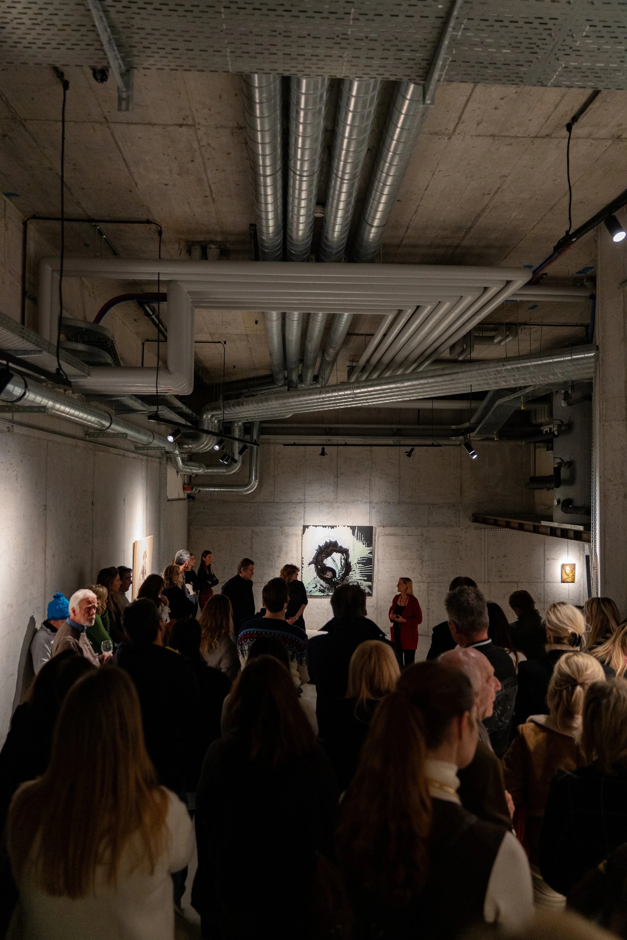 An art gallery or museum exhibit with a group of people gathered around a speaker in front of a painting of a circle or abstract design. The gallery has exposed concrete walls and ceiling with visible pipes and lighting fixtures.