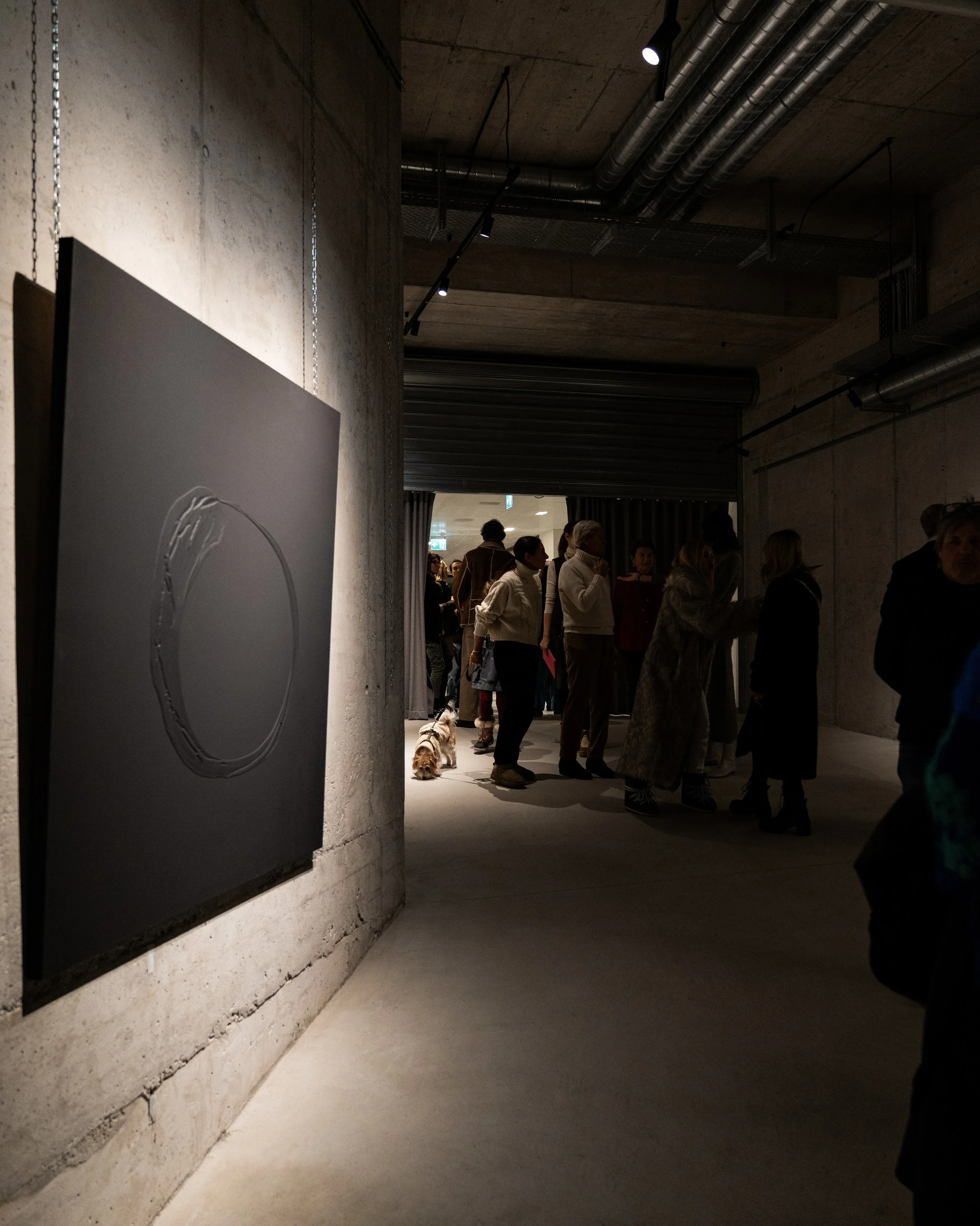 A group of people standing in a room with concrete walls and ceiling, with some people having dogs with them, and artwork hanging on the wall.
