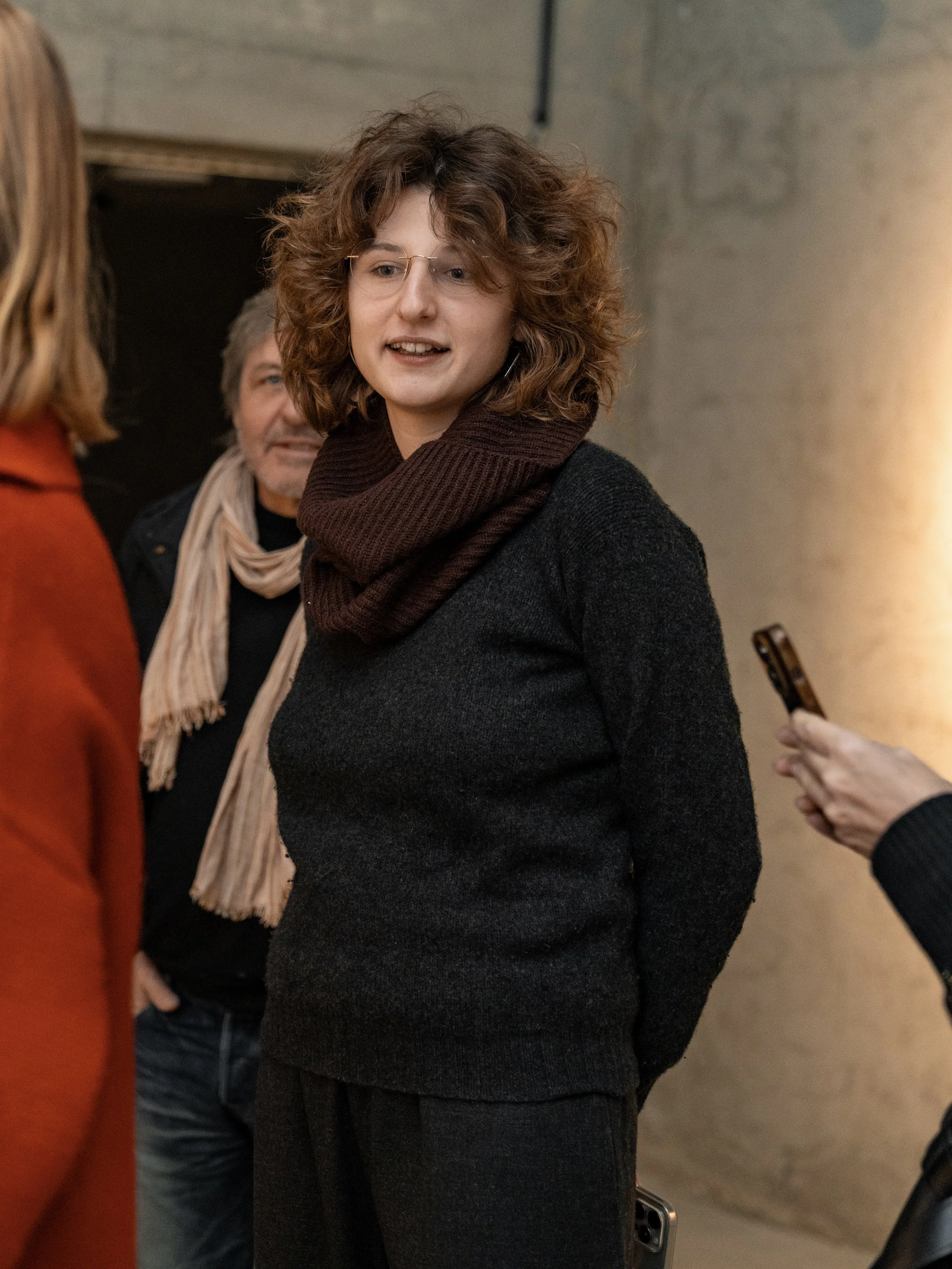 A woman with curly brown hair wearing glasses, a dark sweater, and a brown scarf, talking to a person with shoulder-length hair in an orange top. In the background, a man with gray hair and a beige scarf is visible.