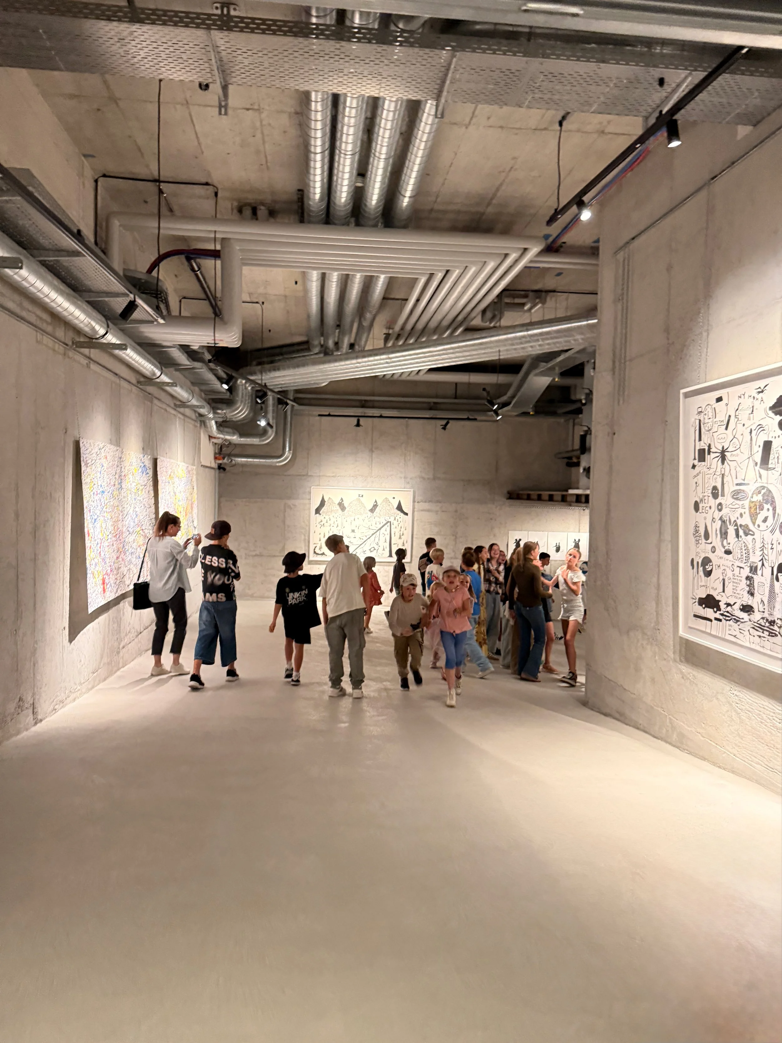 People gathered in an art gallery with concrete walls and ceiling, viewing various artworks displayed on the walls.