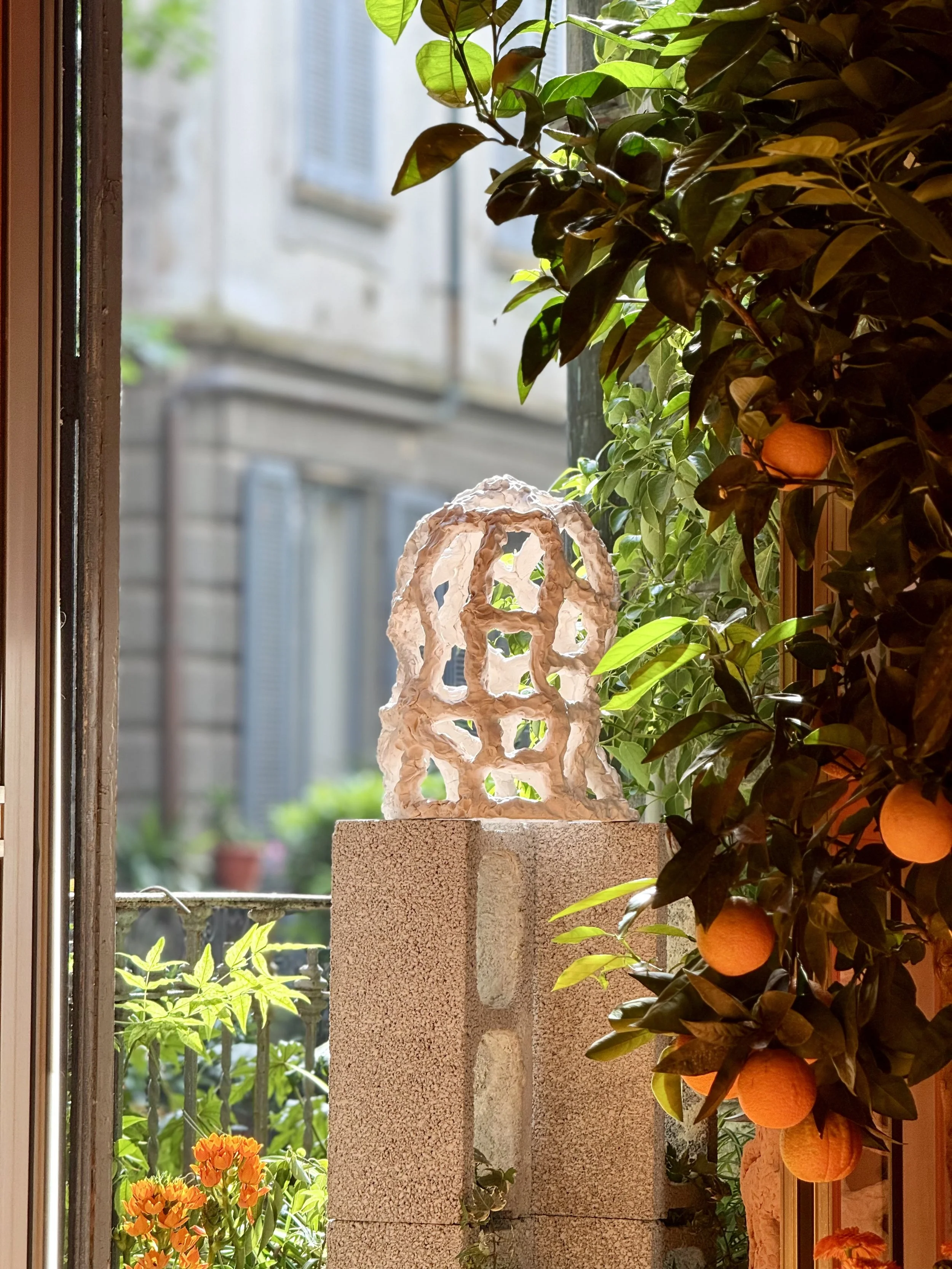 A decorative sculpture made of intertwined white material sits atop a brick pedestal near a window. To the right, a lush orange tree with ripe oranges and dark green leaves is visible. Outside the window, blurred buildings and window shutters can be 