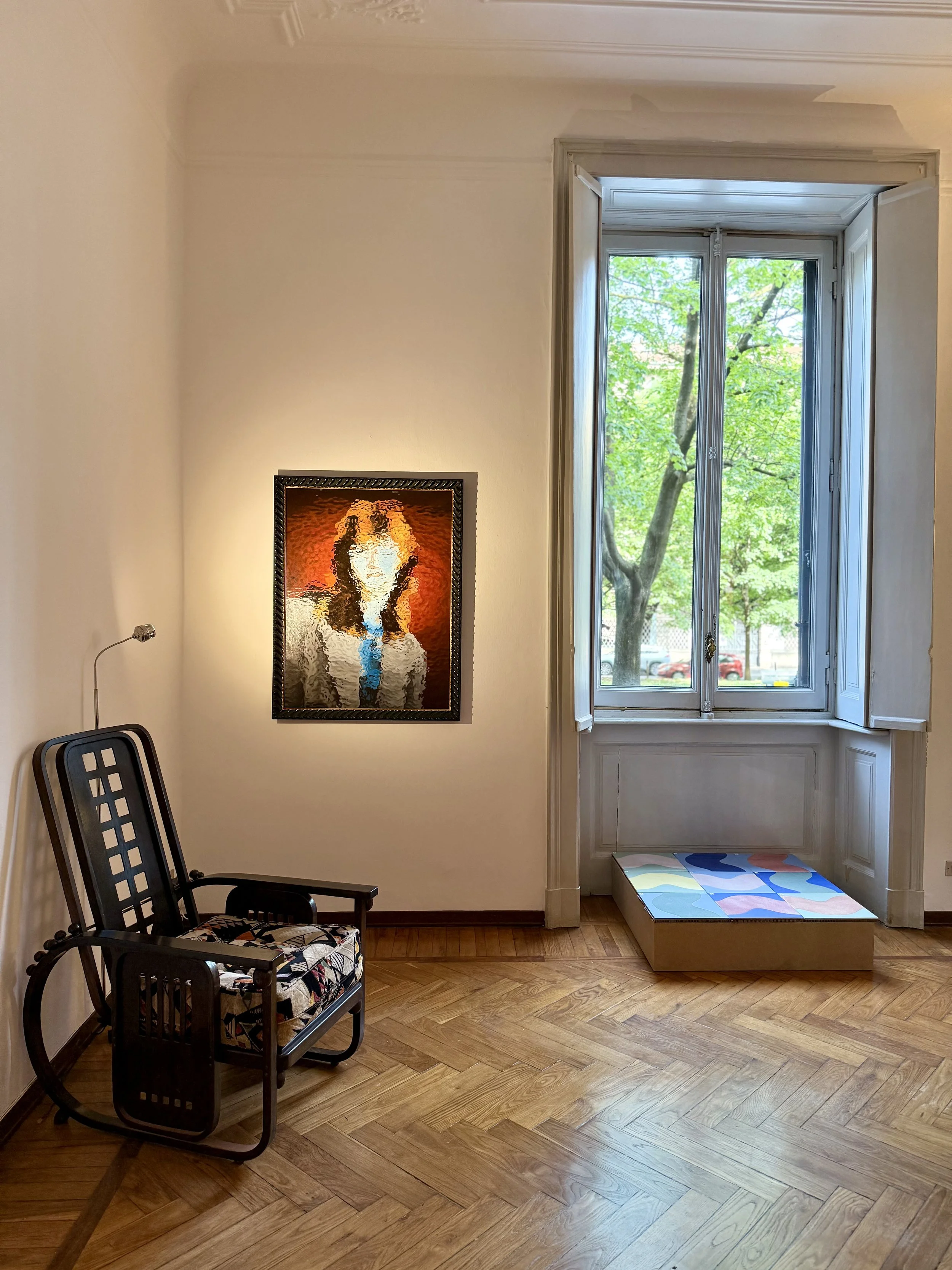 A room with wooden flooring, a large window with a view of green trees outside, a framed portrait of a woman with curly hair on the wall, a black chair with a patterned cushion, a bench with a colorful abstract design, and a small lamp on the chair