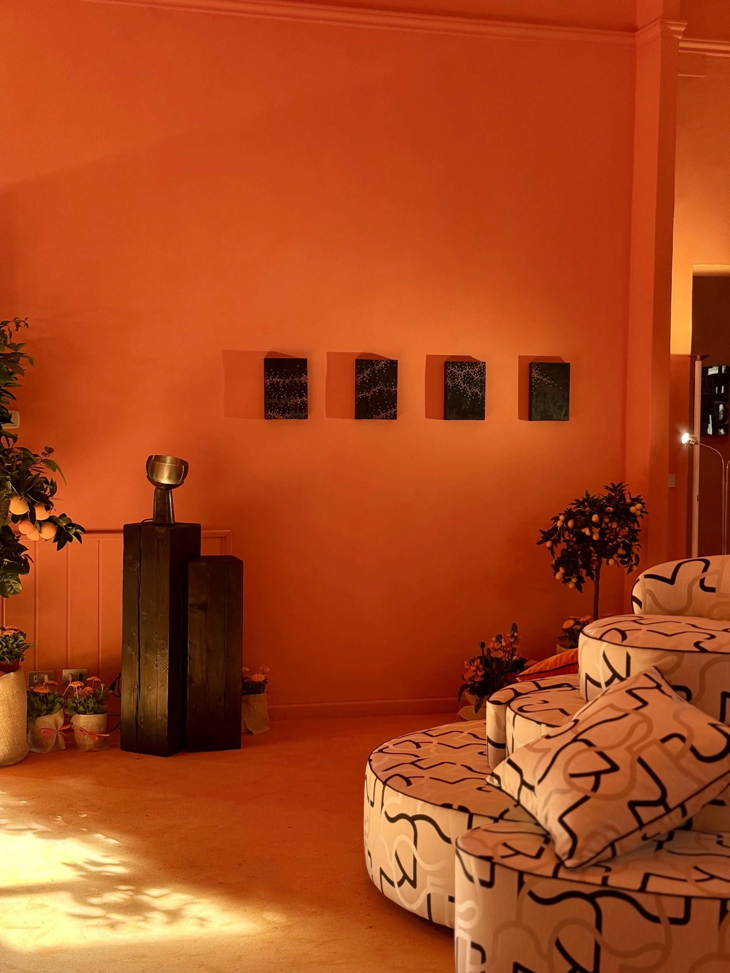 A cozy living room with a large orange wall decorated with four black framed artworks. There is a white sofa with black abstract patterns and a rolled-up blanket, a small potted plant with yellow flowers, and some smaller potted plants on the floor. The room is illuminated by warm lighting, casting shadows on the wall and floor.