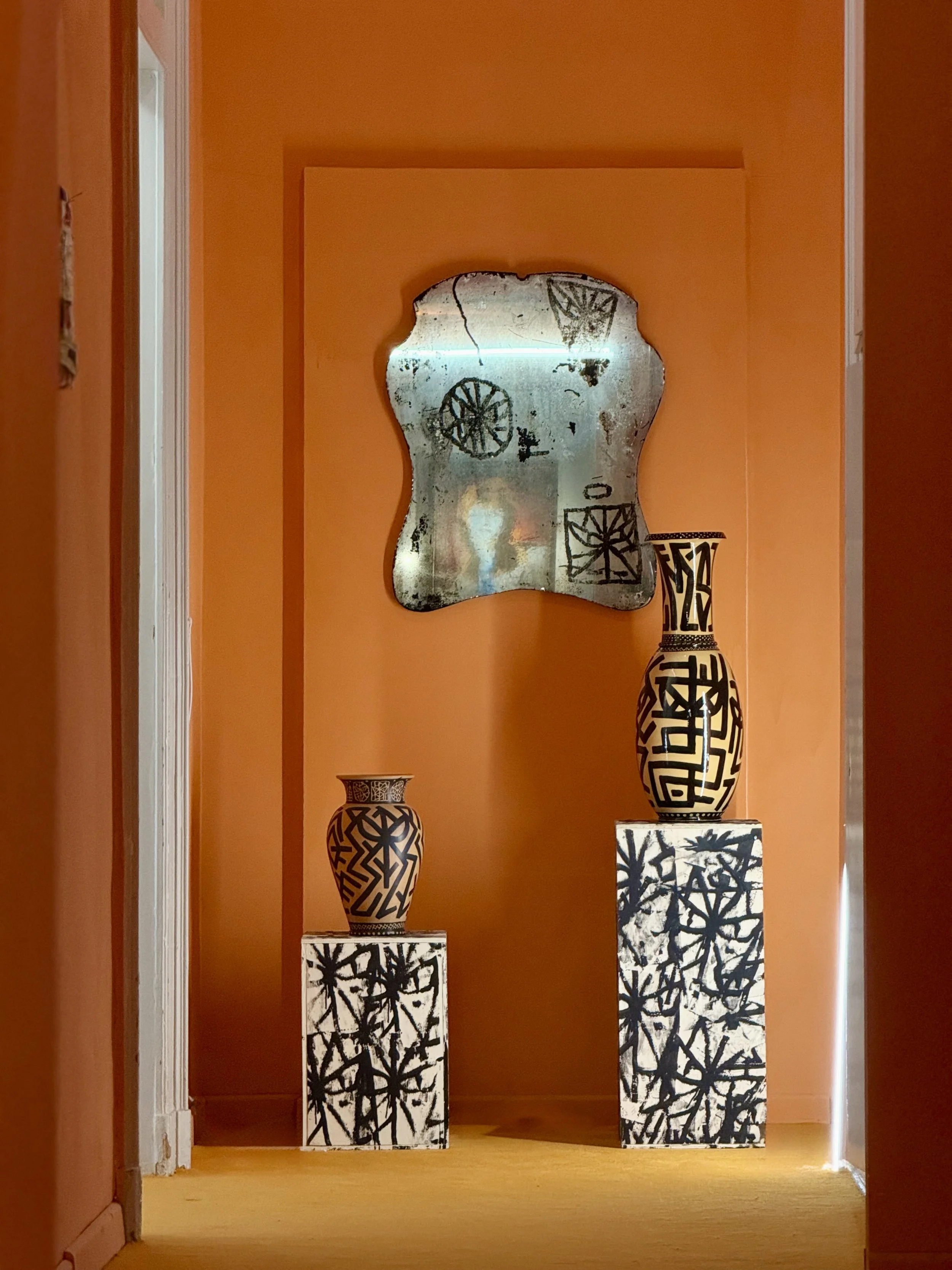 A display of black and white patterned vases and sculptures on white pedestals against an orange wall with a decorative mirror.