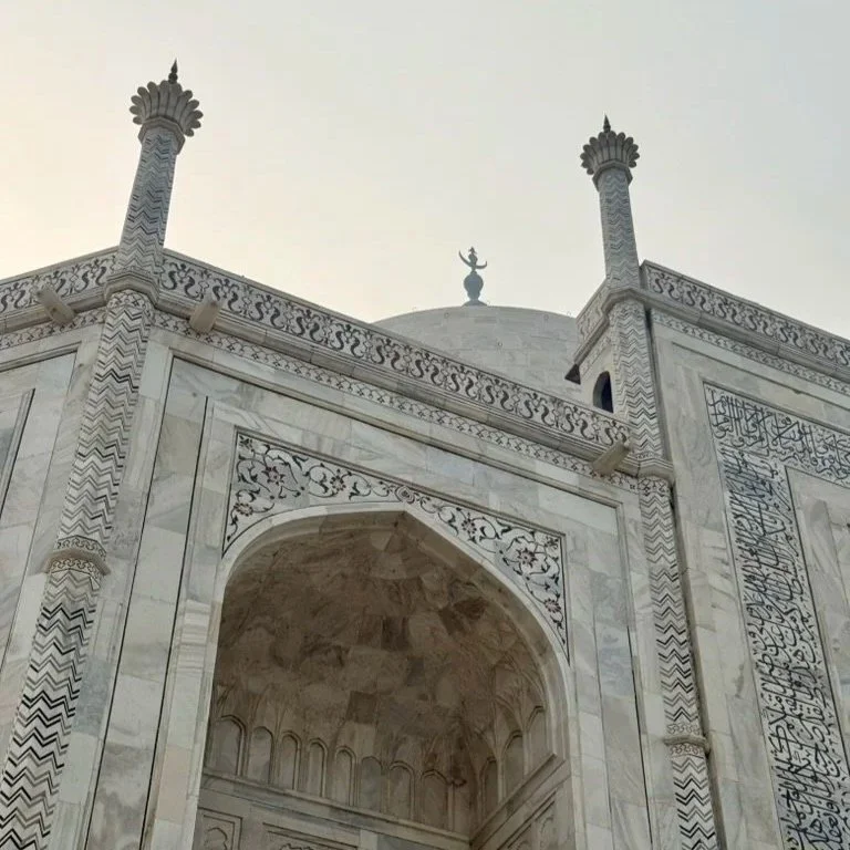 A close-up view of the marble Mughal-style architecture of the Taj Mahal, showing ornate arches, minarets, and a dome with a finial on top. The sky is clear.