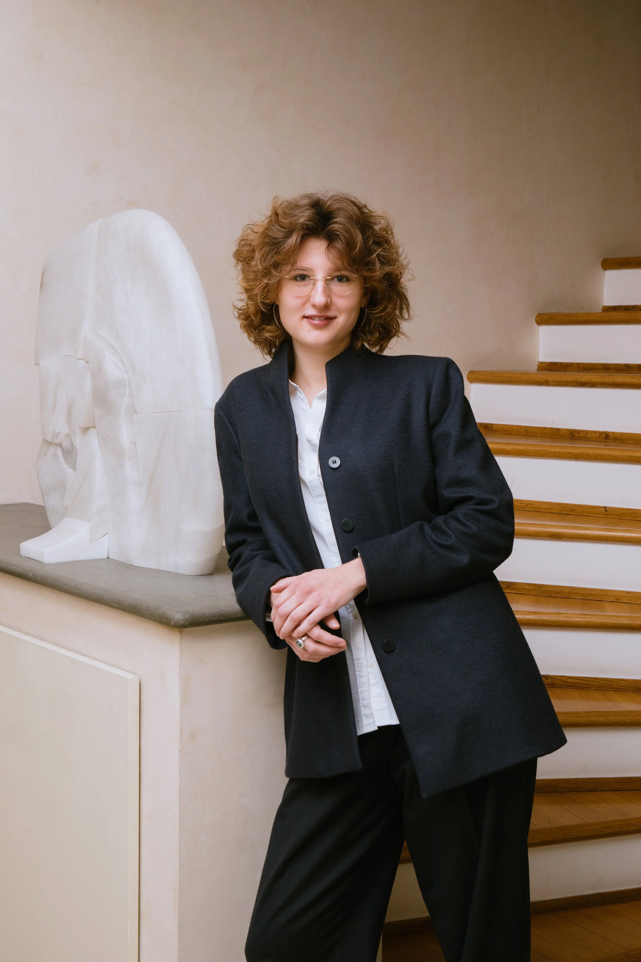 A woman with curly hair and glasses wearing a dark blazer and white shirt, standing indoors near a staircase, with a white marble sculpture of an angel in the background.