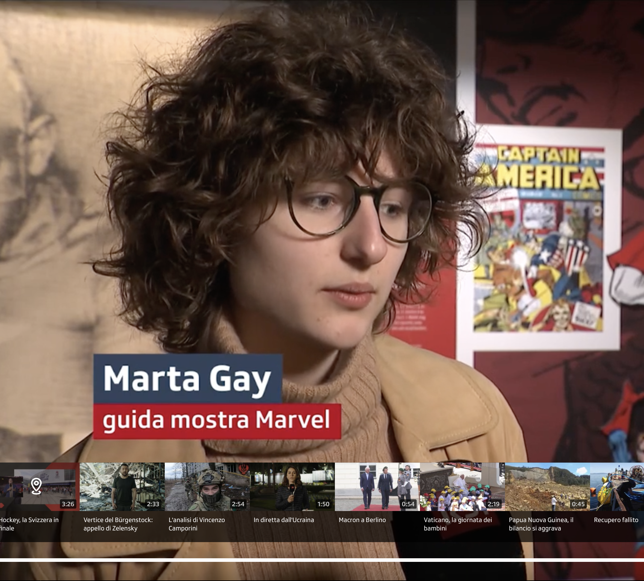 Close-up of a woman with curly brown hair and glasses, wearing a tan sweater, in front of a mural and a poster of Captain America.