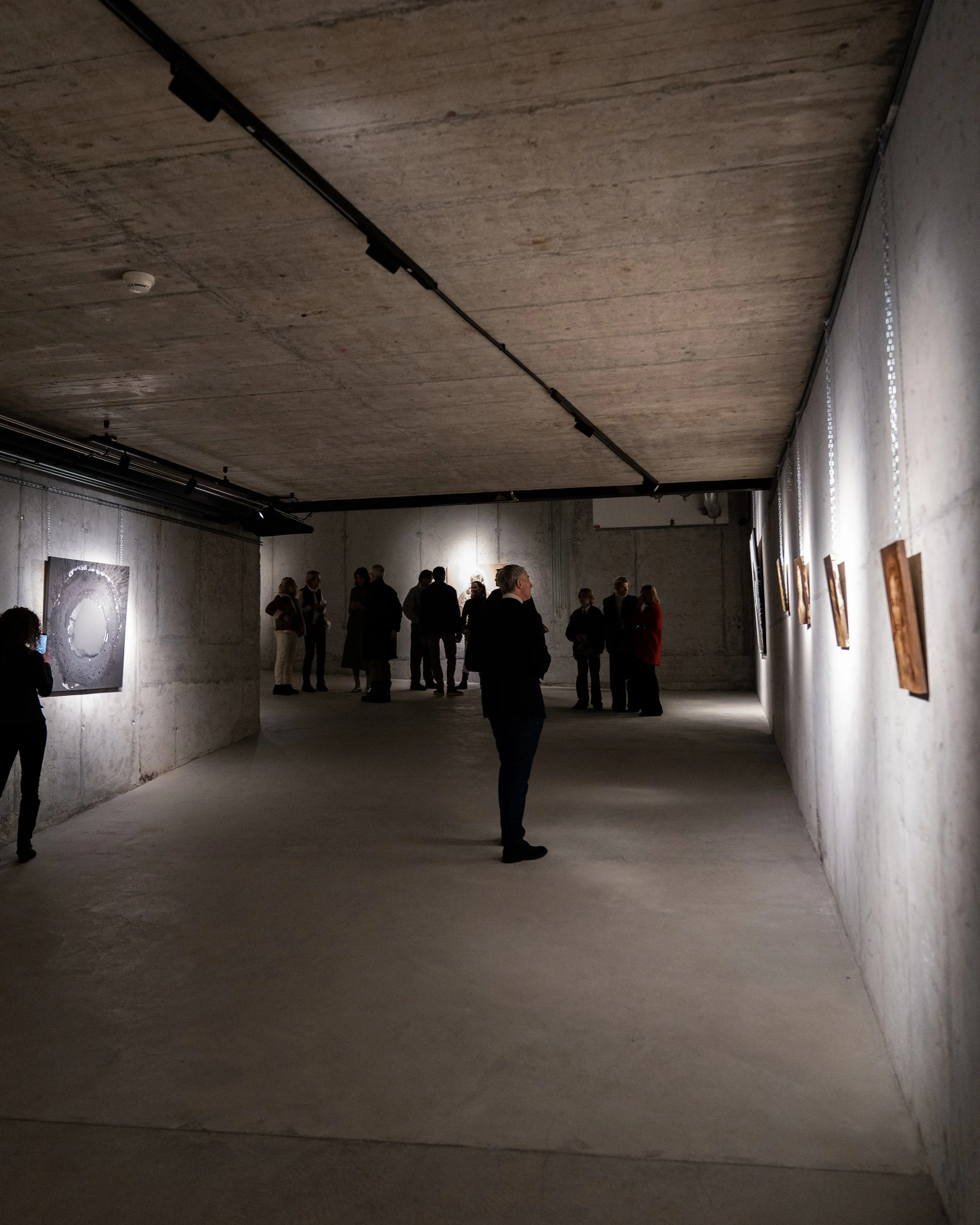 An art gallery with concrete walls and ceiling, featuring artworks on display and visitors observing them, with a group of people gathered and conversing.