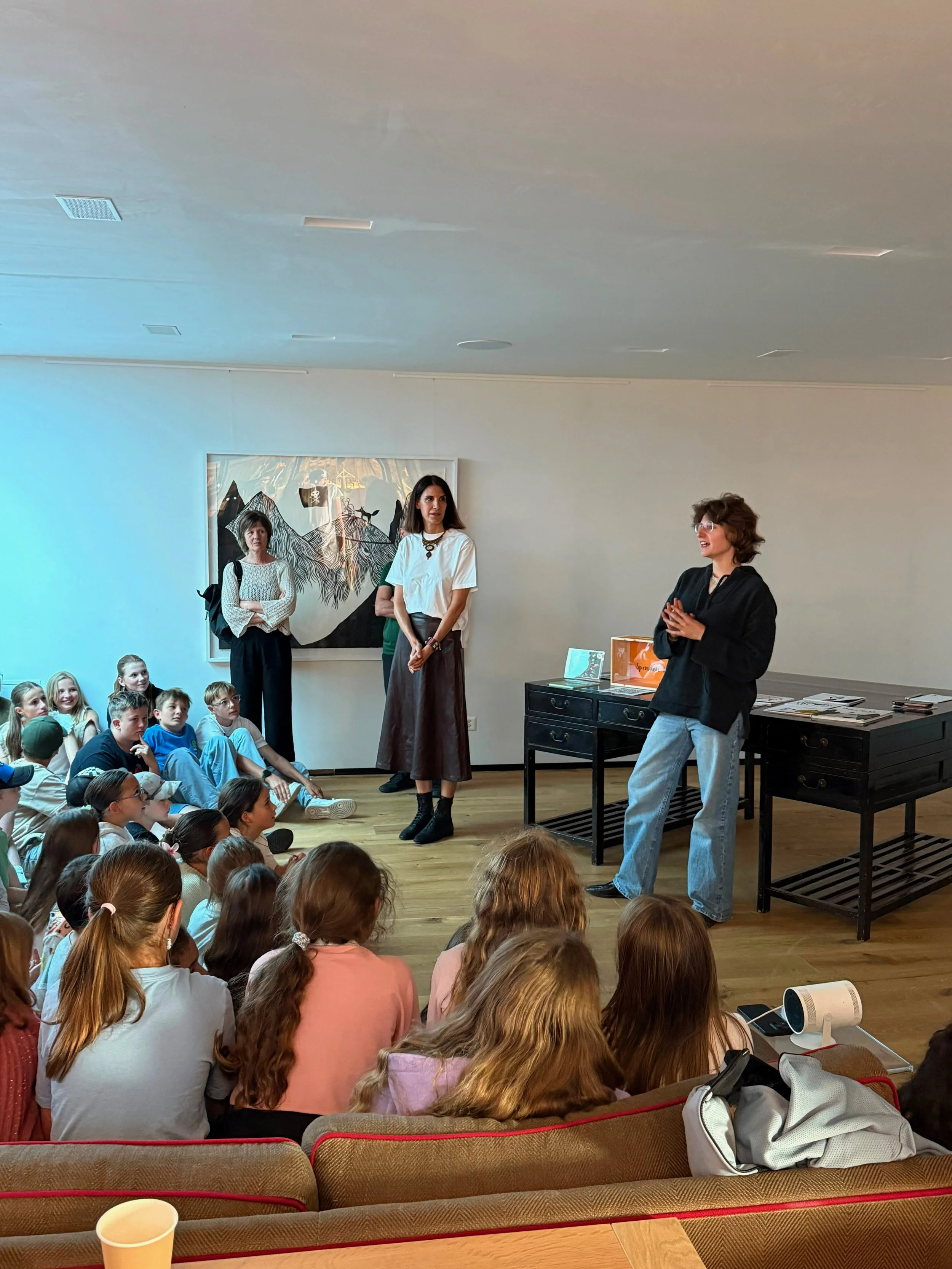 A group of children sitting on the floor watching a presentation or discussion led by three women standing near a wall with artwork. One woman is talking and smiling, another woman is standing with arms crossed, and the third woman looks on. The room has a wooden floor and a black table with books or materials.