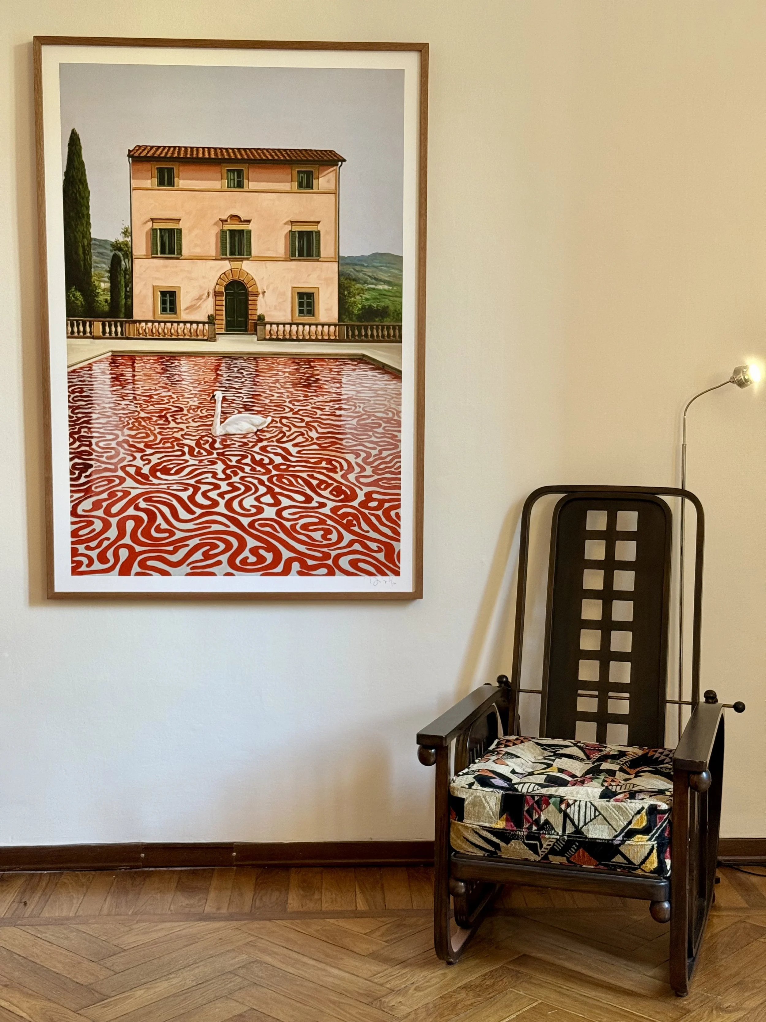 A framed painting of a pink Italian villa with a red tile roof and a courtyard with a red and white abstract pattern, a white swan in the center, surrounded by greenery and distant hills, hangs on a beige wall. Next to a dark wooden armchair with col