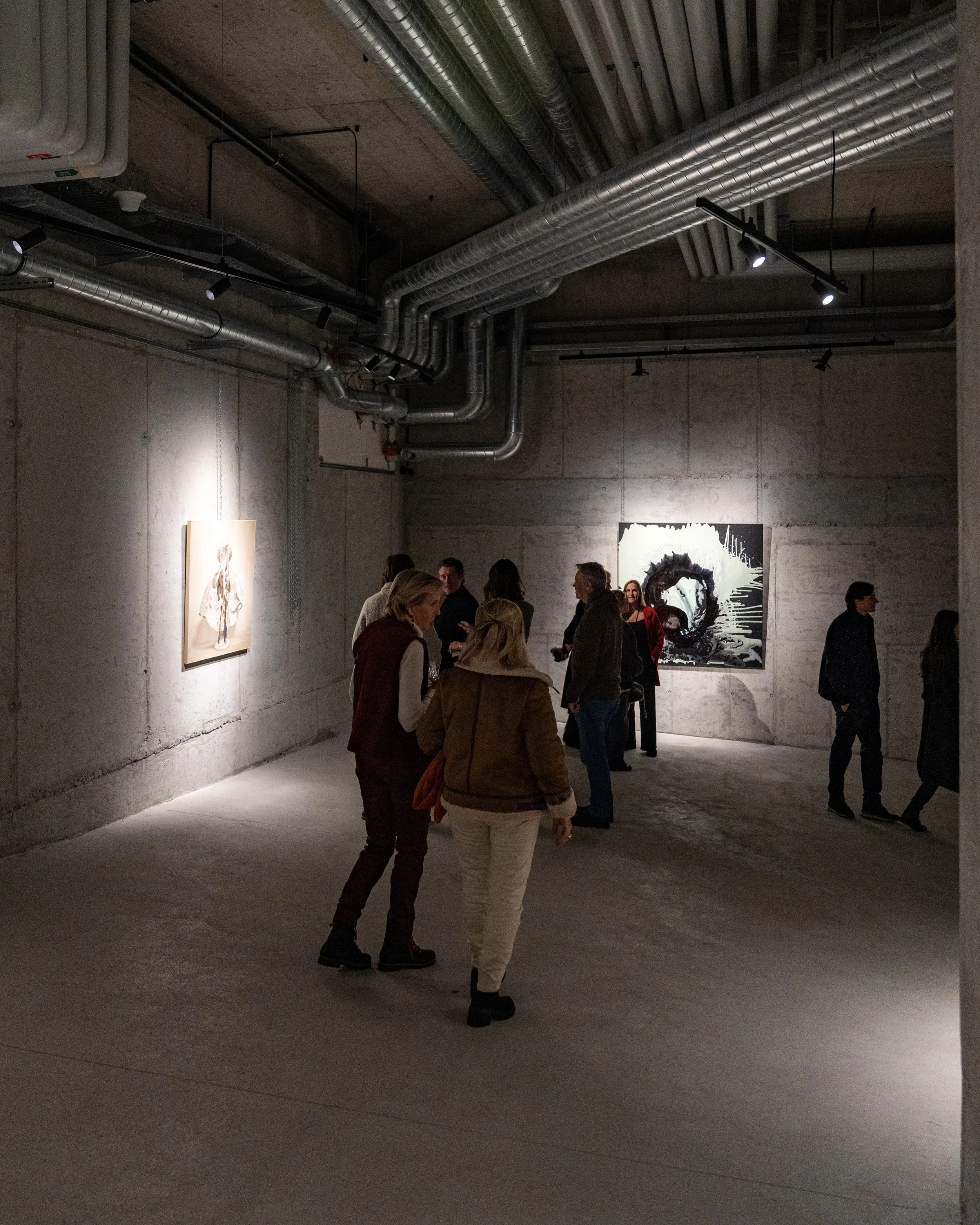 People gathered in an art gallery or museum, viewing artwork on the wall, with industrial-style exposed pipes and concrete walls
