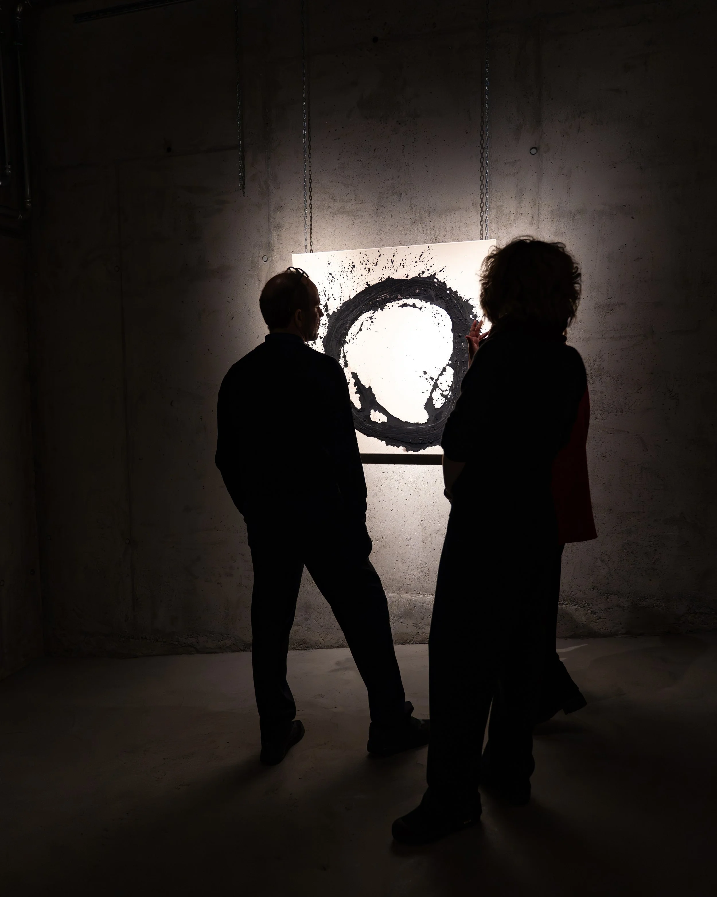 Two people having a conversation in front of a large monochrome portrait of a man's face at an art gallery or exhibition with concrete walls.