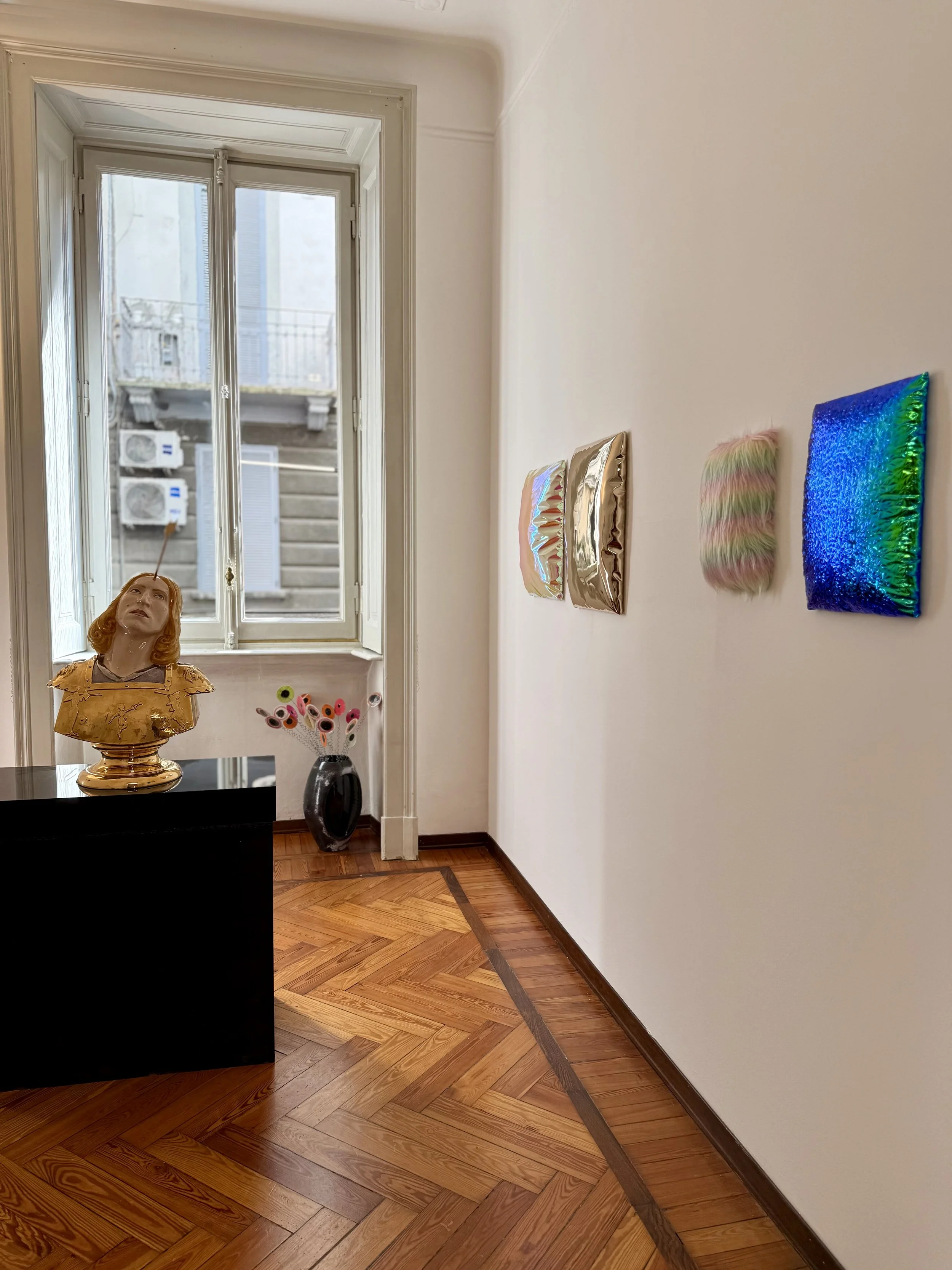 A corner of an art gallery with three textured, colorful art pieces on a white wall, a wooden floor, a mannequin bust with vintage clothing on a black pedestal, a window with a city view, and a vase with colorful artificial eyes.