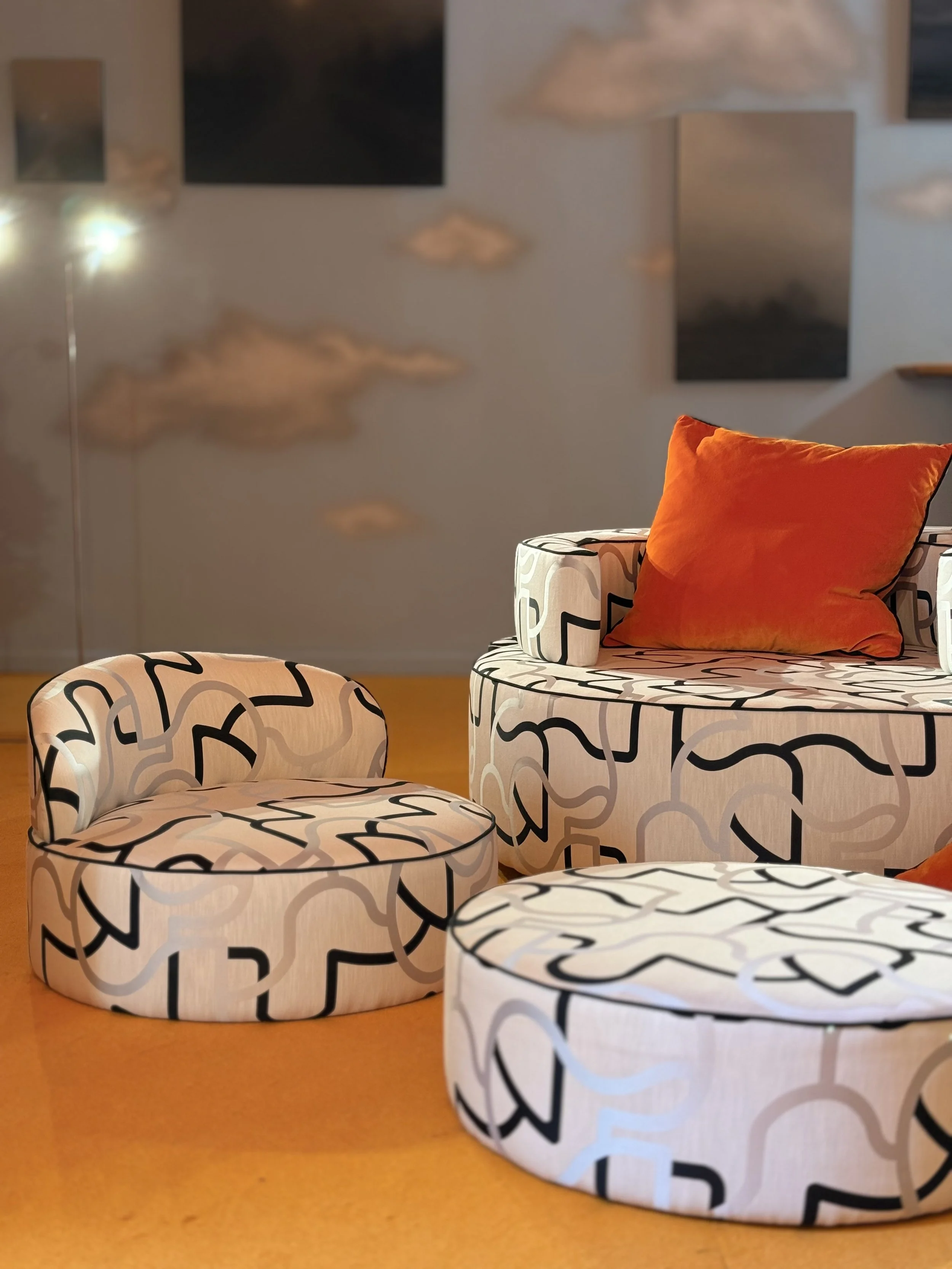 A room with furniture covered in white fabric with black abstract patterns, including ottomans, a chair, and a sofa. The room has a white wall decorated with abstract paintings and hanging cloud-shaped light fixtures, with a warm-colored carpet on the floor.