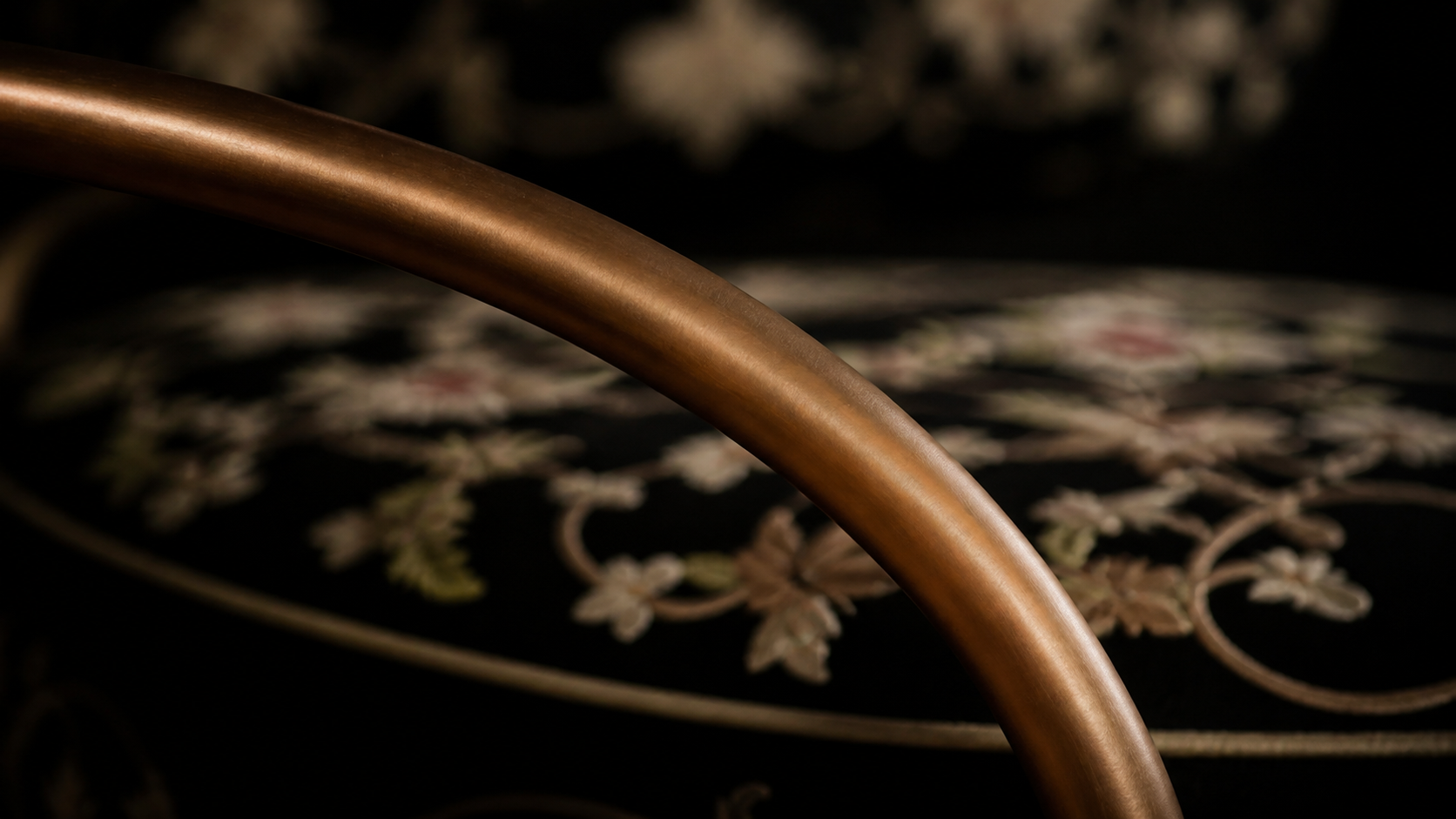 Close-up of a curved copper-colored metal frame in front of a decorative floral embroidery on a dark fabric background.