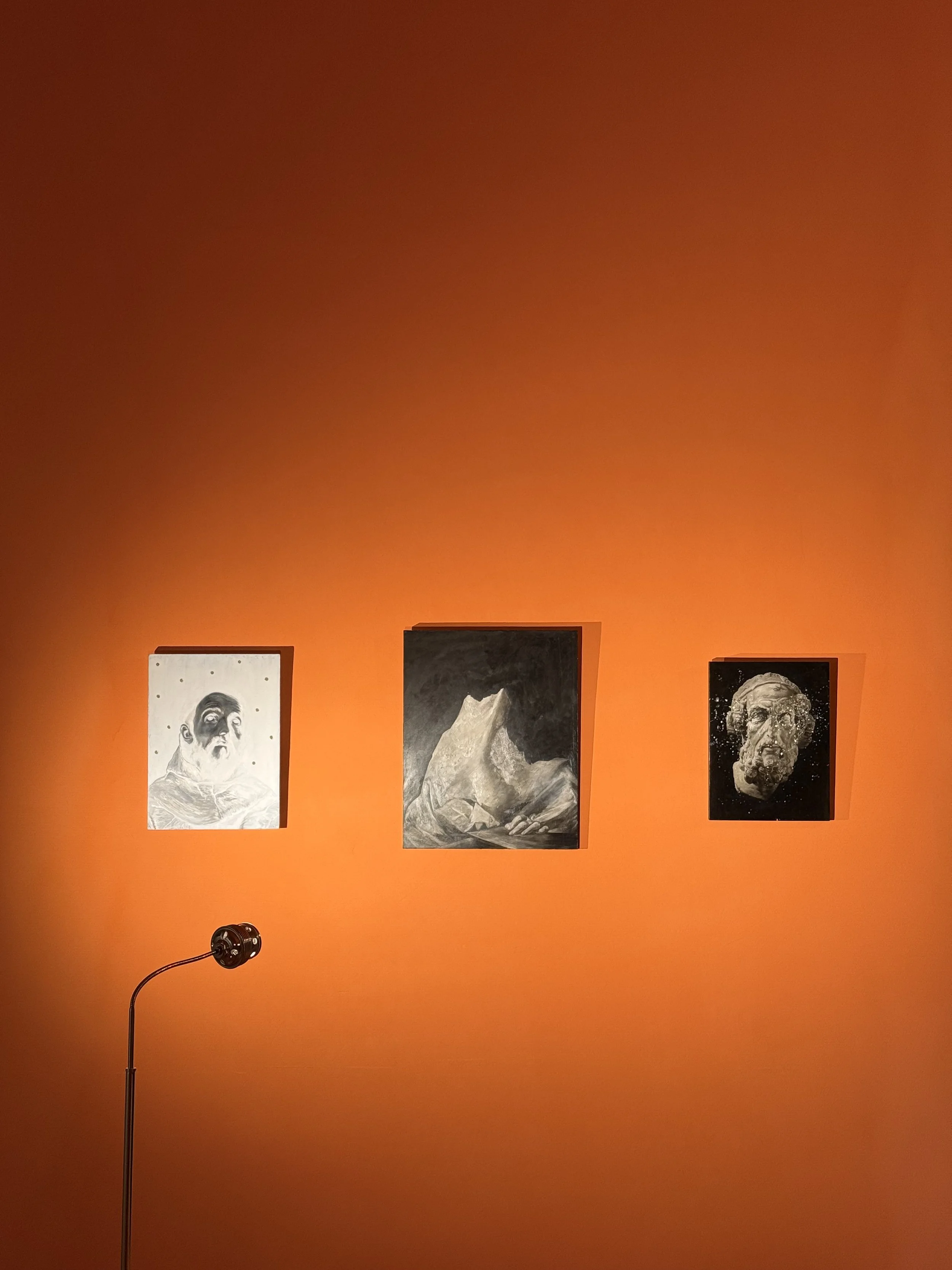Three black and white art prints are hung on a burnt orange wall, with a small black lamp or projector standing on a stand in front of them.