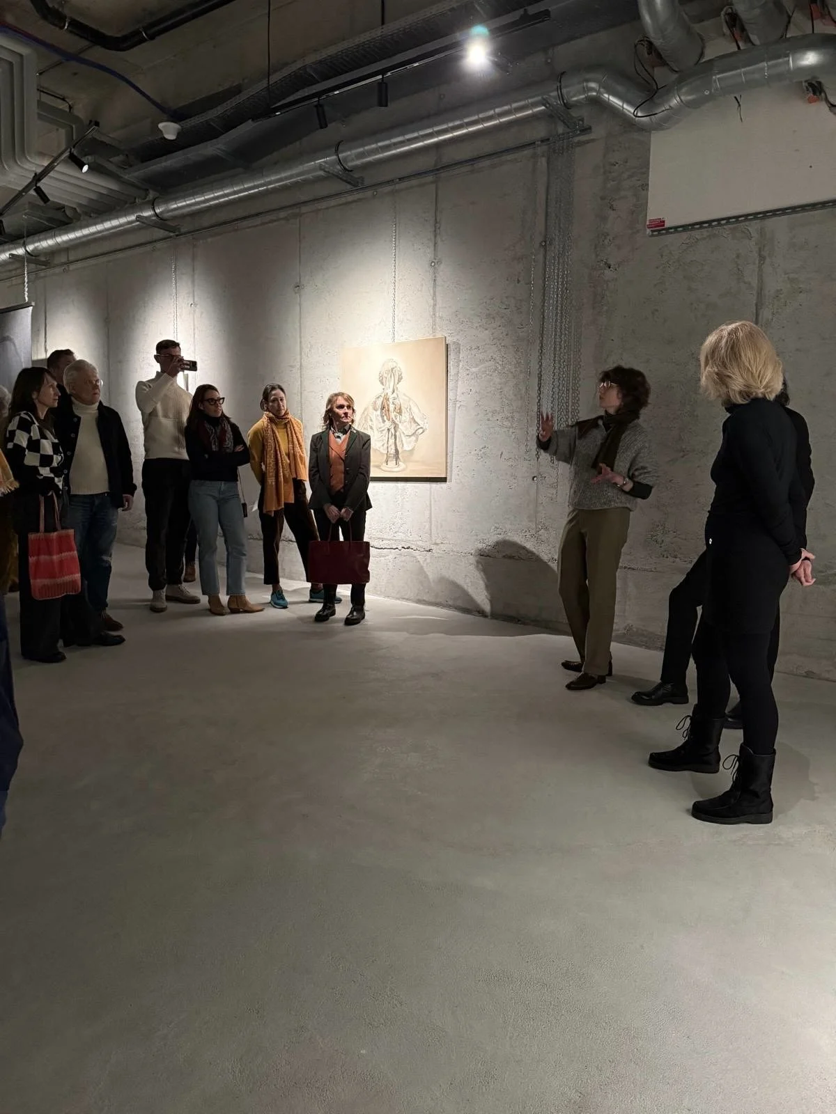 A group of people attending an art gallery or museum exhibit, listening to a woman explaining artwork on a concrete wall.