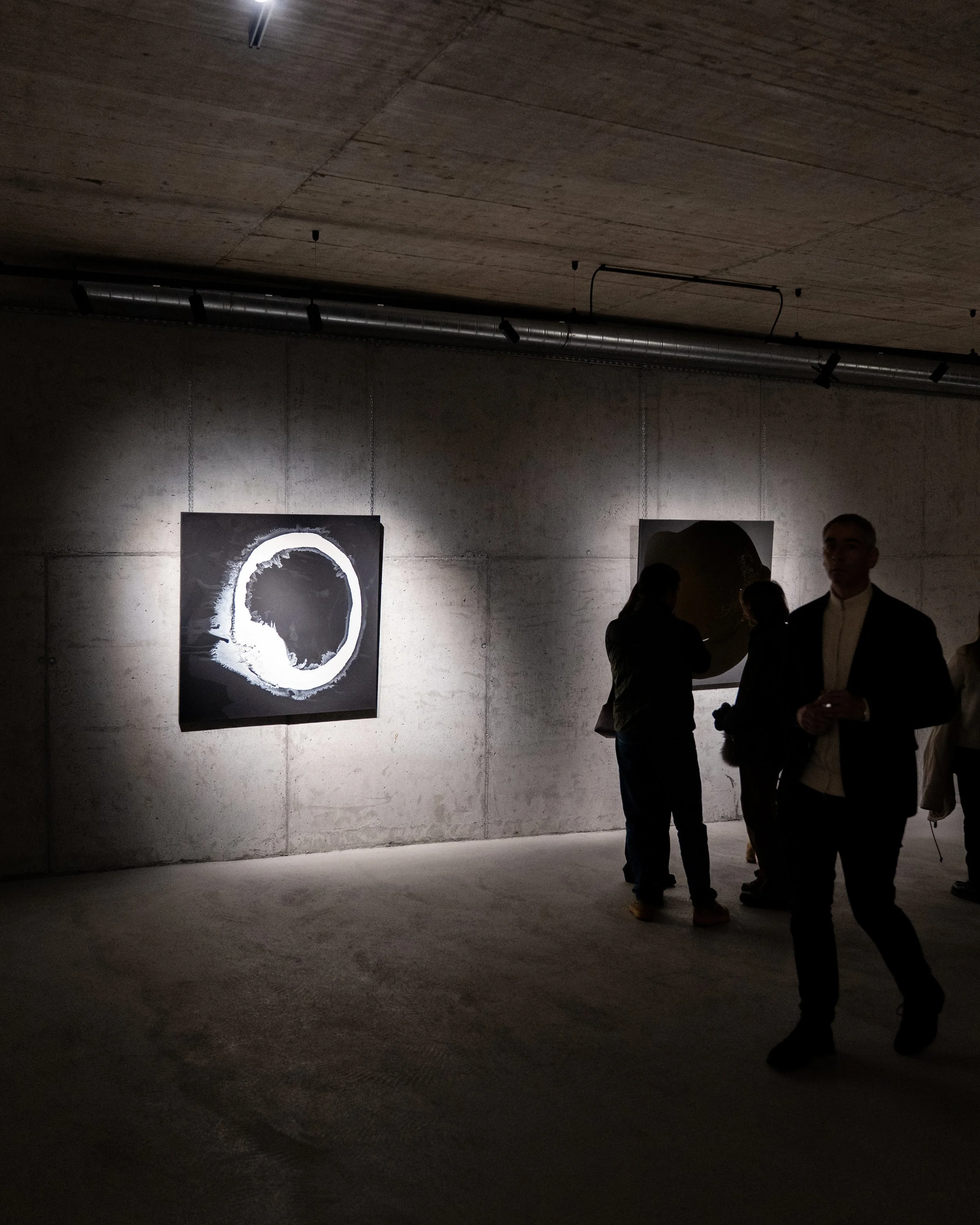 An art gallery with dark lighting, featuring a large black and white circular art piece hanging on a concrete wall, and several people standing and conversing in the background.