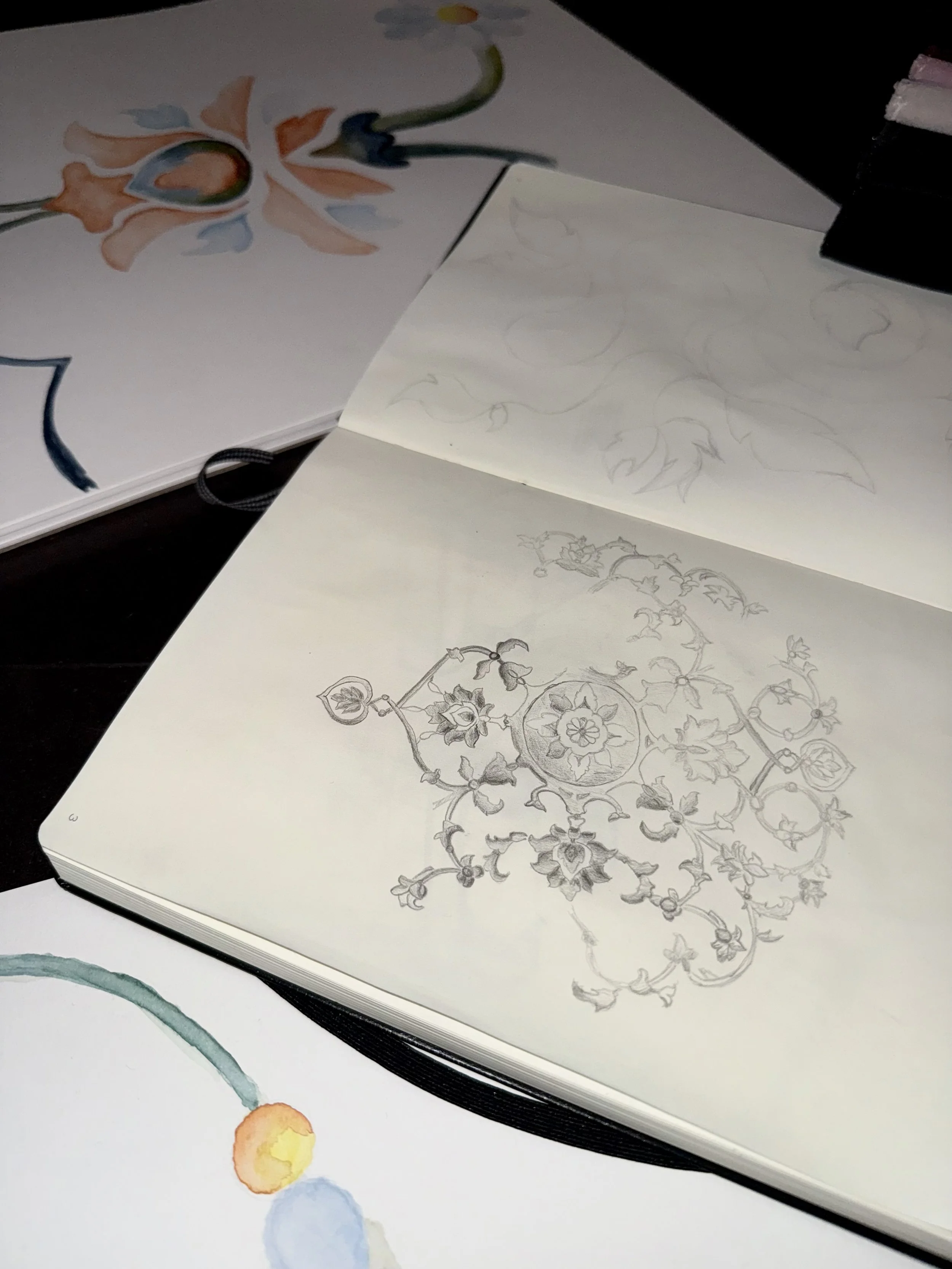 A sketch of intricate floral and vine patterns on paper, alongside a painted floral design with pastel colors.