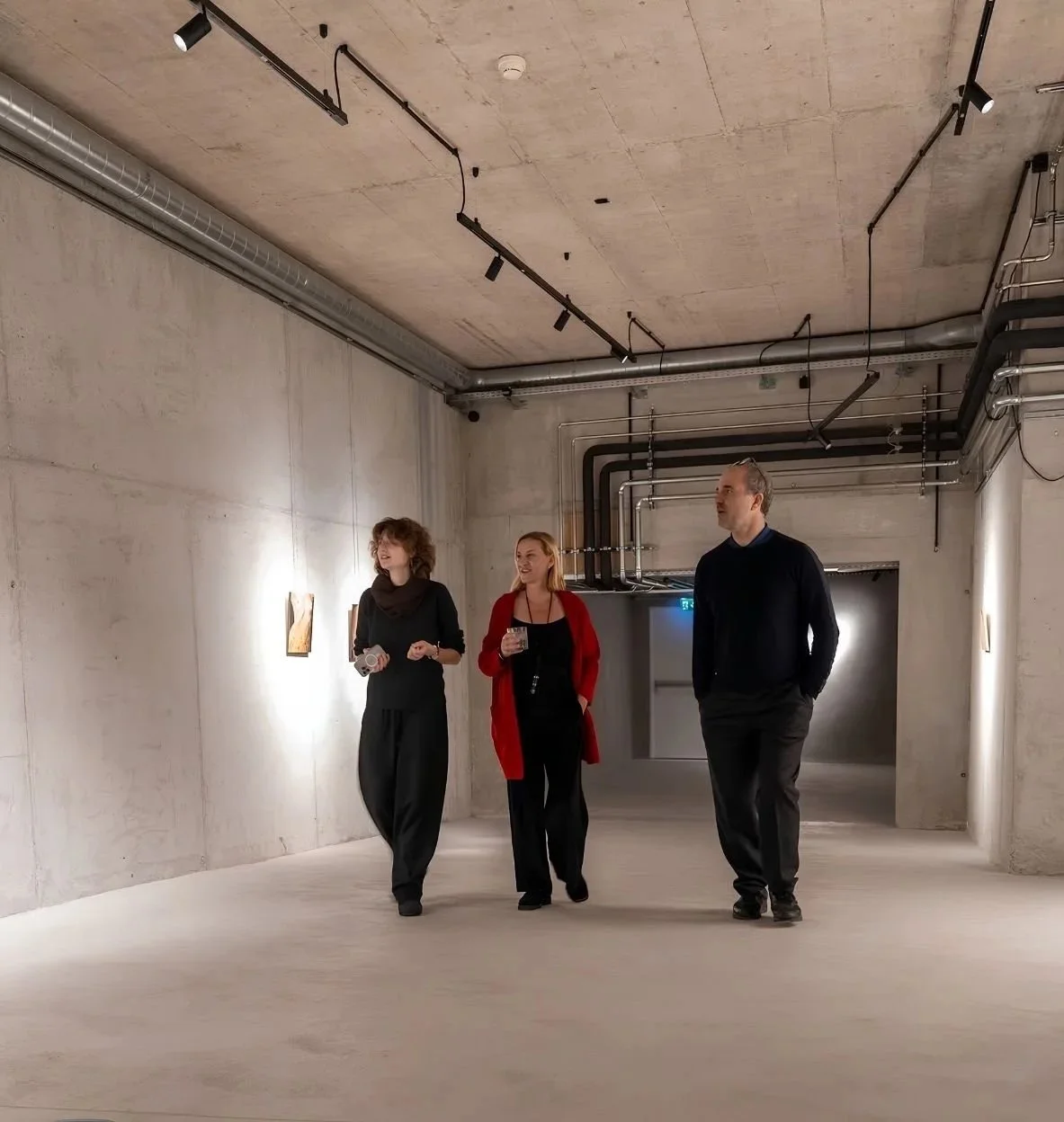 Three people walking and talking in an art gallery with minimalist concrete walls and ceiling, featuring exposed pipes and track lighting.